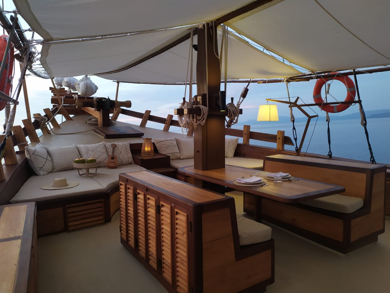 Warmly lit foredeck of Senja at blue hour with cushioned lounge, ukulele and set dining table