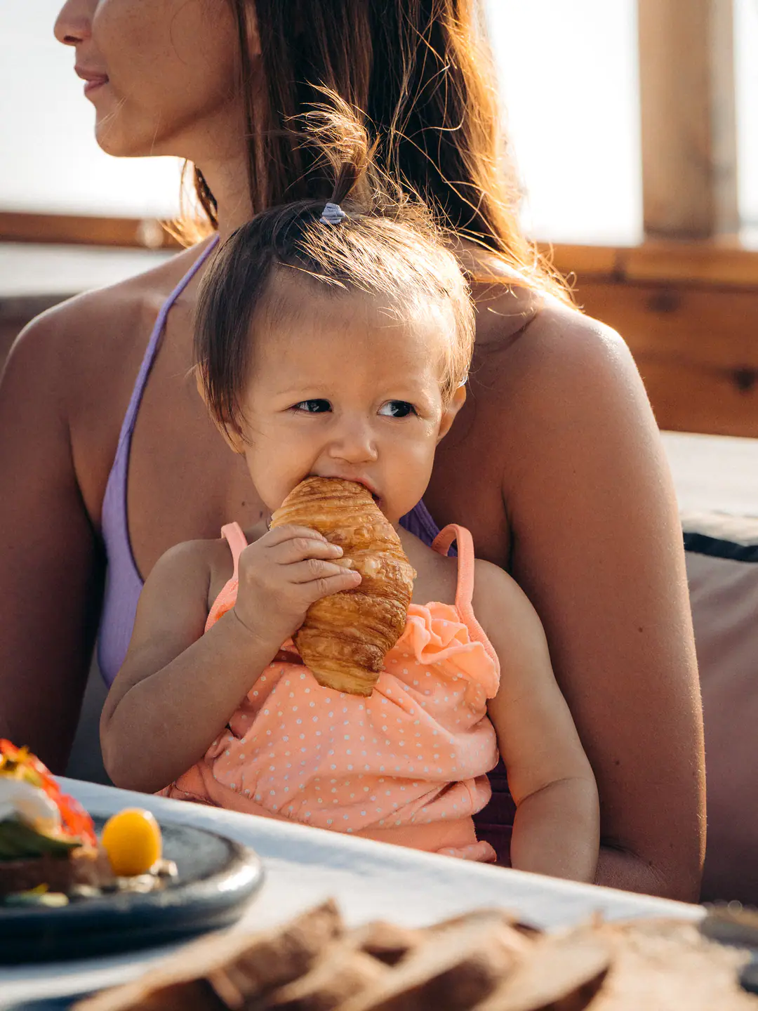 Toddler on mother's lap biting into a croissant in warm morning light on The Oracle