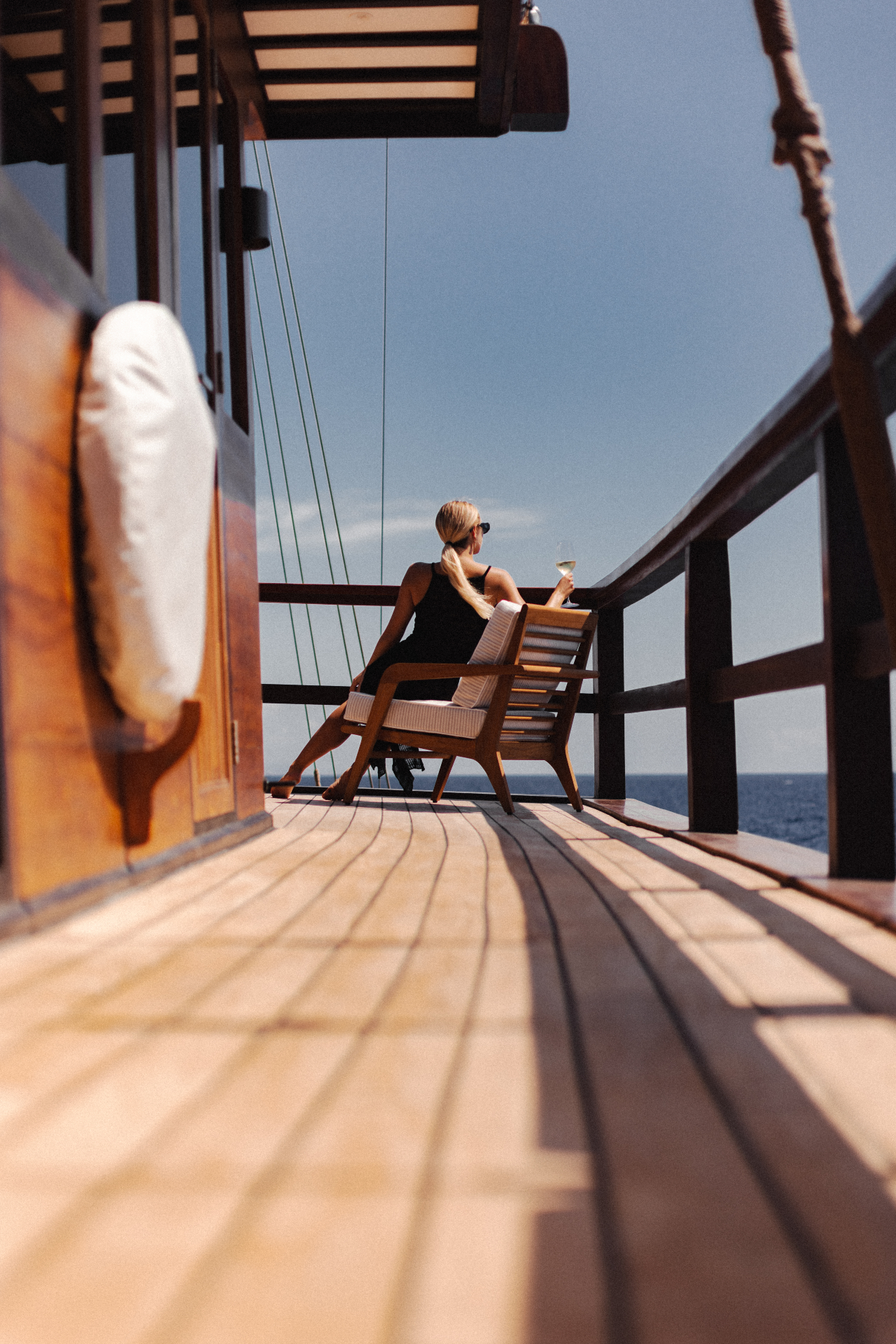 Guest seated on Vela's side deck at midday with a glass of white wine and open ocean beyond the rail