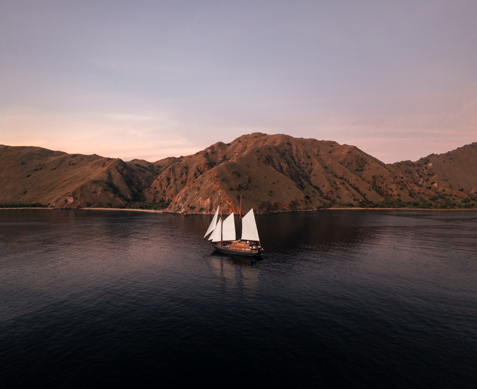 Vela under full white sail on calm water at sunset with arid Komodo hills behind