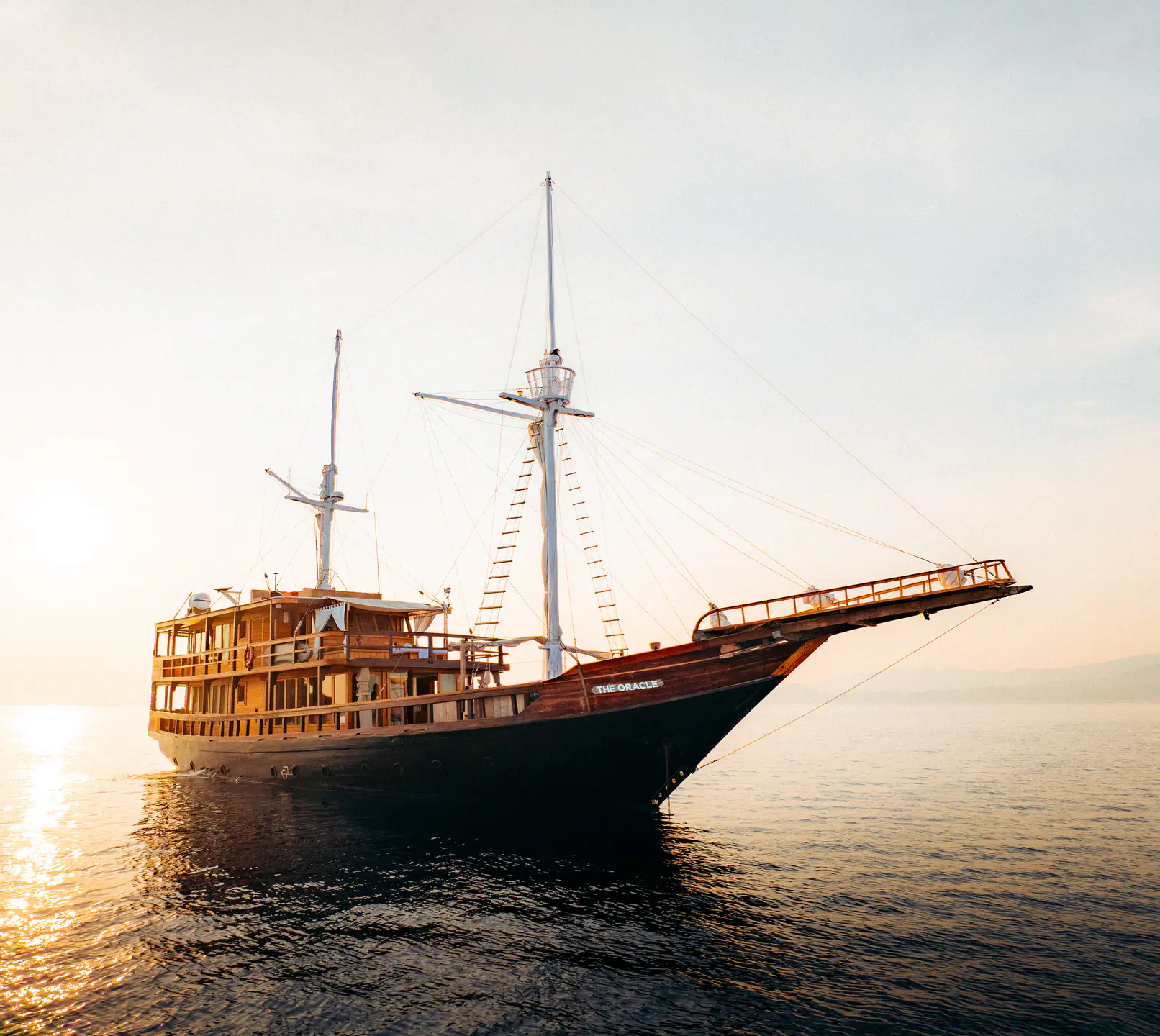The Oracle phinisi at anchor in golden sunset light with two masts silhouetted