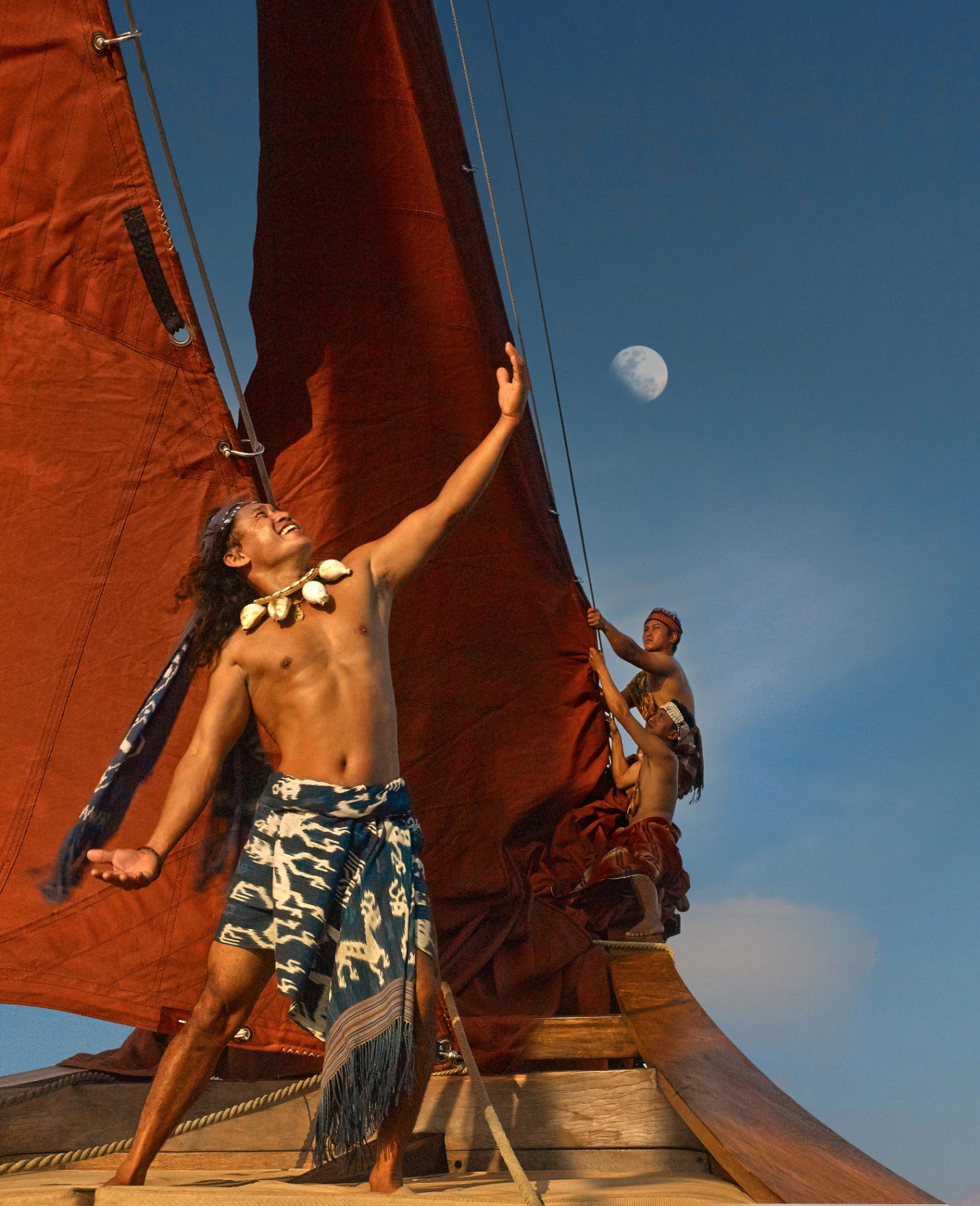 Si Datu Bua crew in traditional ikat sarongs hoisting the crimson gaff sails at moonrise