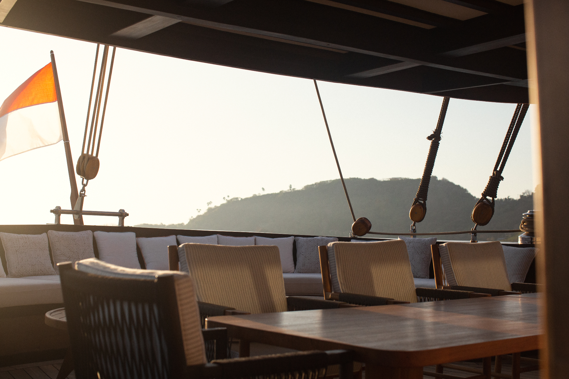 Aft deck seating on Vela at golden hour with Indonesian flag, rope blocks and hills in soft backlight
