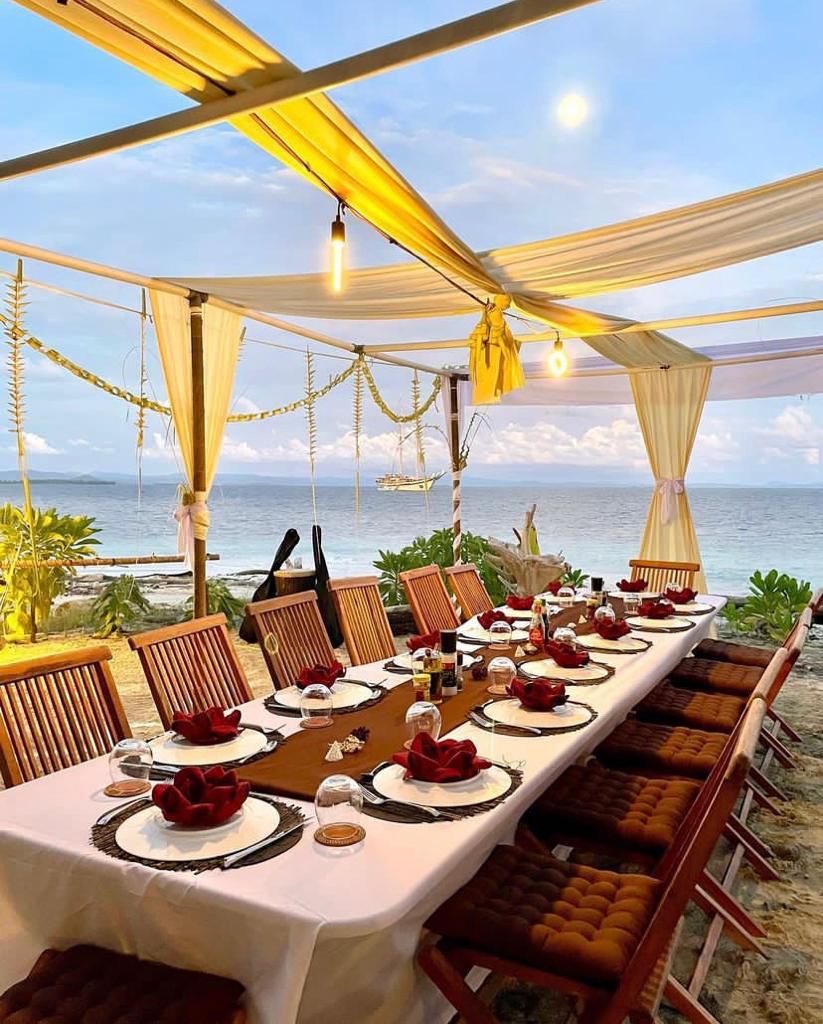 Beachside dinner setup on a Raja Ampat sandbank with Lamima anchored offshore at sunset