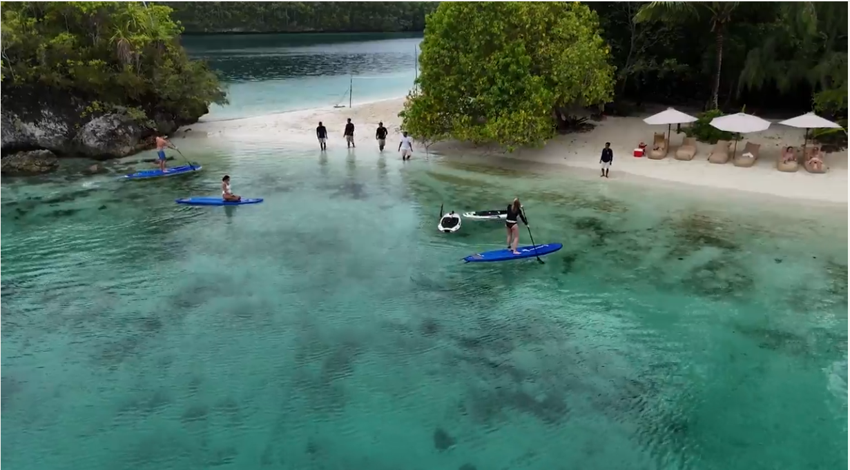 Guests stand-up paddleboarding and kayaking on a shallow turquoise lagoon beside a private beach setup with parasols