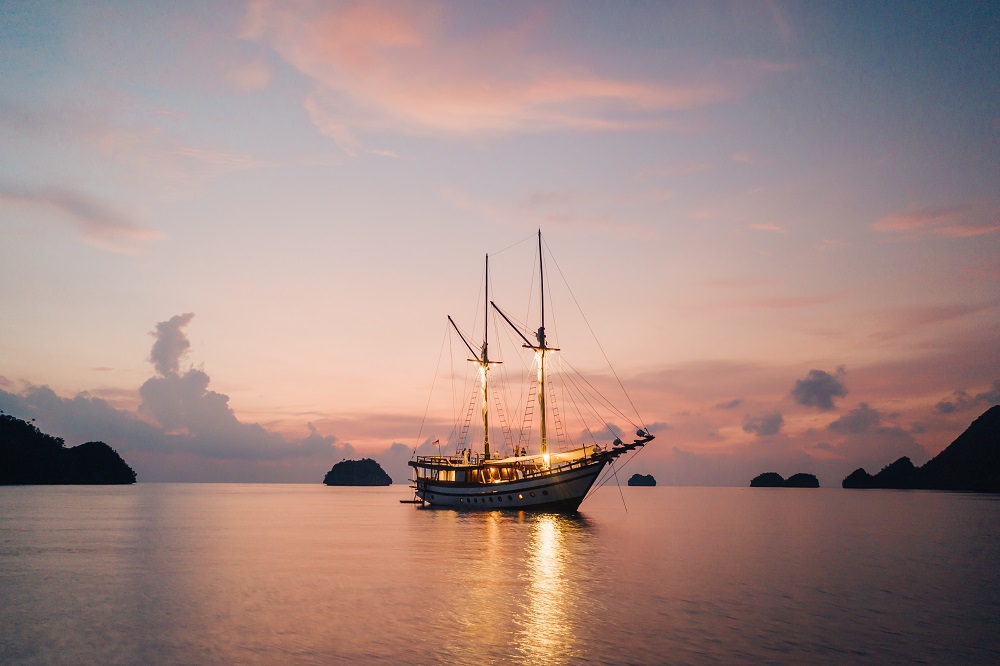 Senja lit up at anchor beneath a pink and purple Raja Ampat sunset with karst islands around