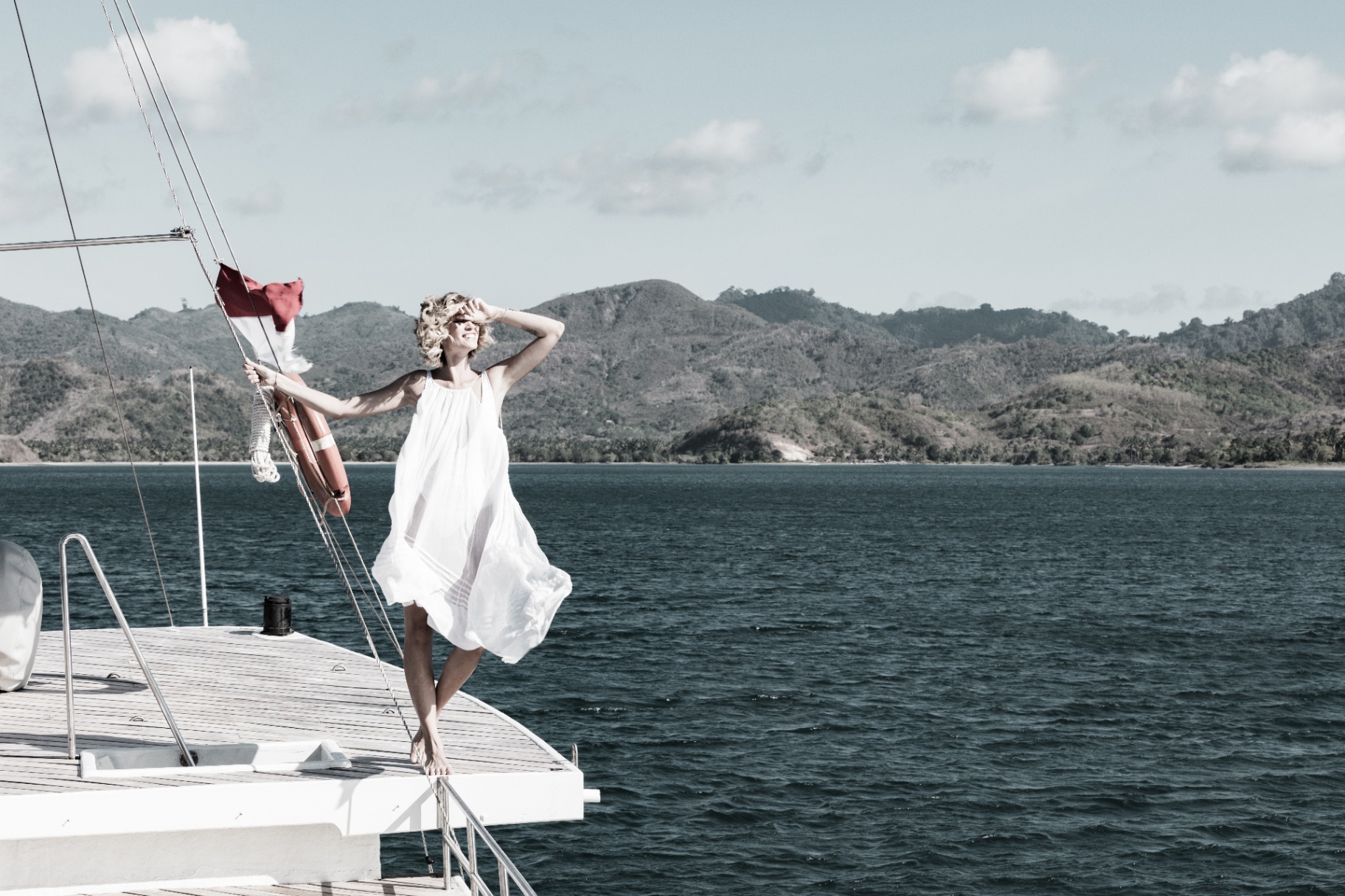Woman in a flowing white dress standing on Splendour's bow with mountains and Indonesian flag behind