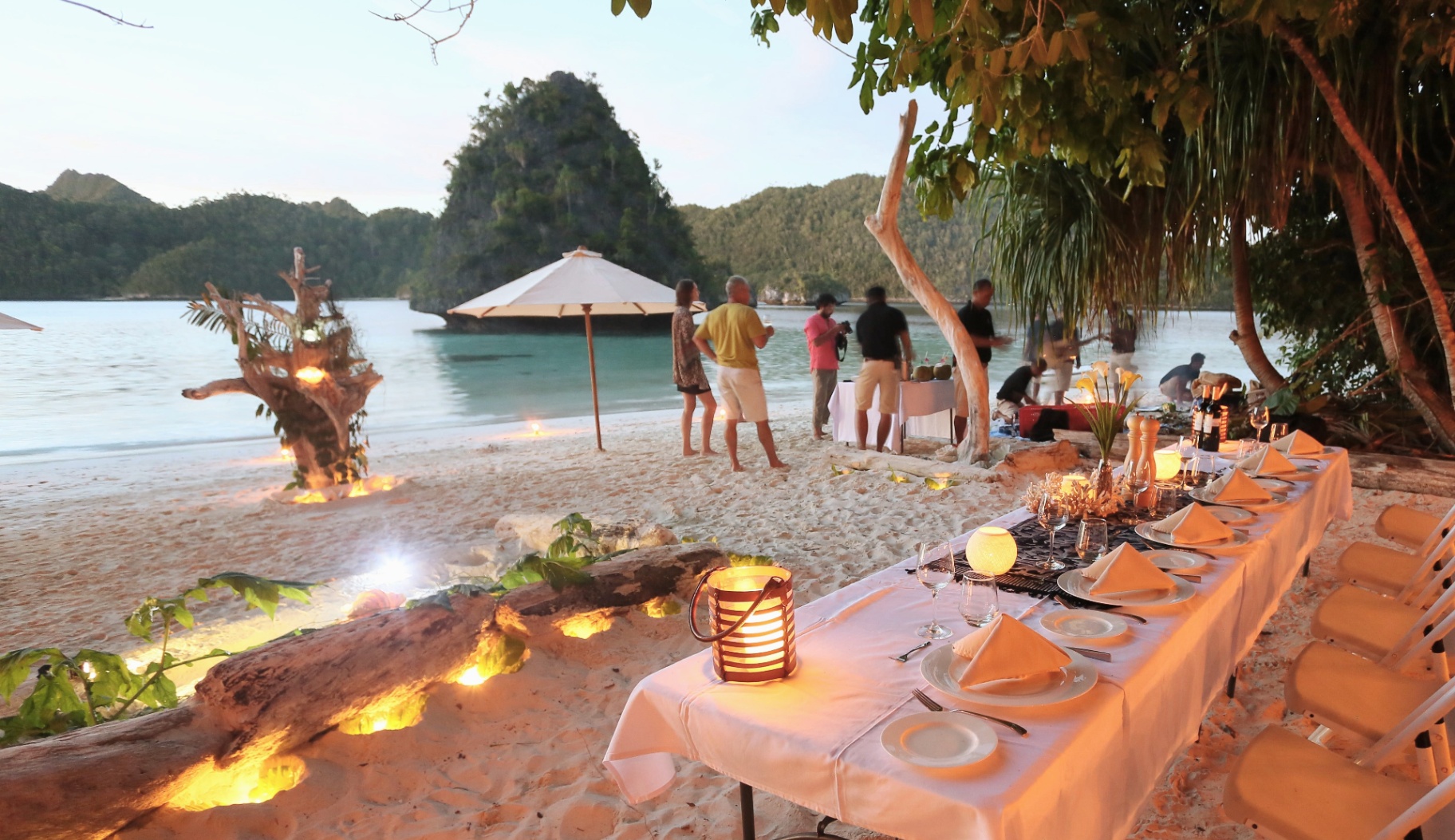 Beach barbecue table set with candles and driftwood in front of a lone Wayag karst pinnacle at golden hour