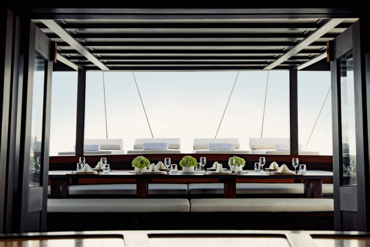 Sky-deck dining table set beside white sun daybeds under a slatted canopy