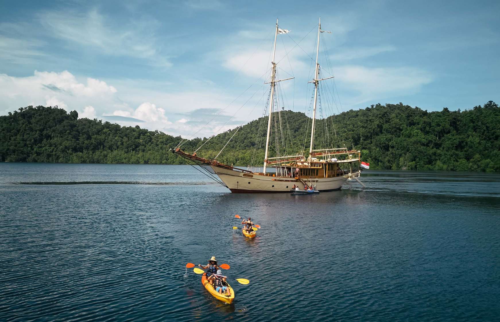 Si Datu Bua anchored in a lush green bay with two yellow kayaks paddling in the foreground