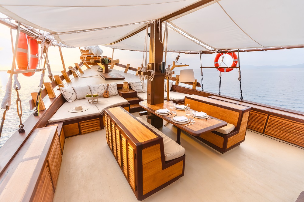 Al fresco dining set on Senja's foredeck beside the cushioned bow lounge, framed by white sun sails and open sea