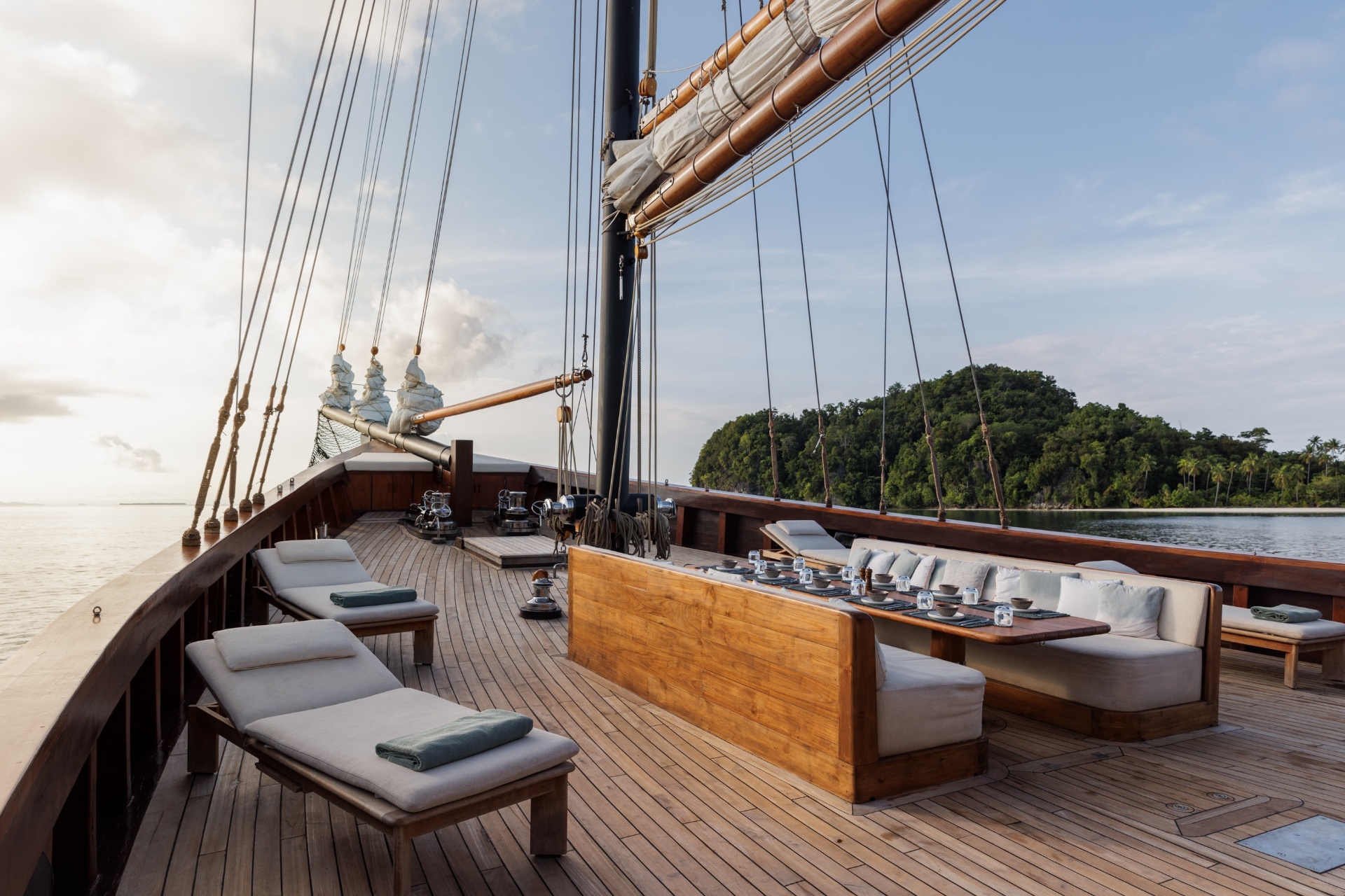 Vela's foredeck at golden hour with teak sunloungers, a long dining banquette set for service and twin masts overhead