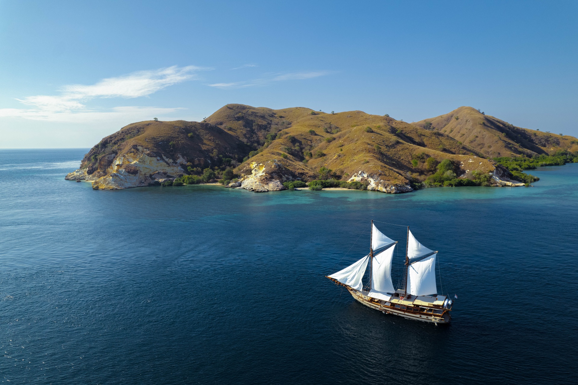Nataraja sailing under full white sails past a green Komodo island on deep blue water