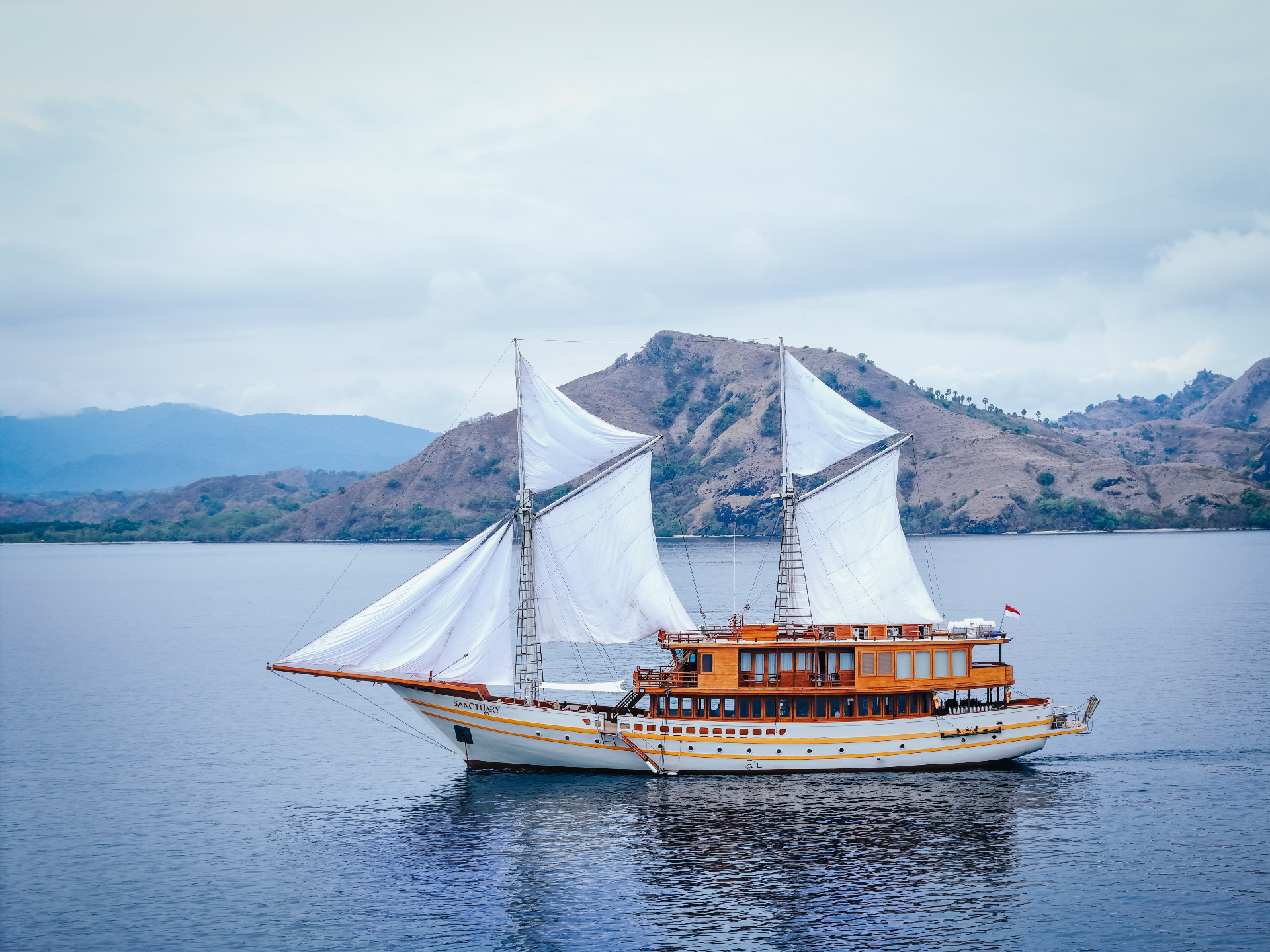 Sanctuary sailing under full white sails past the dry hills of Komodo National Park