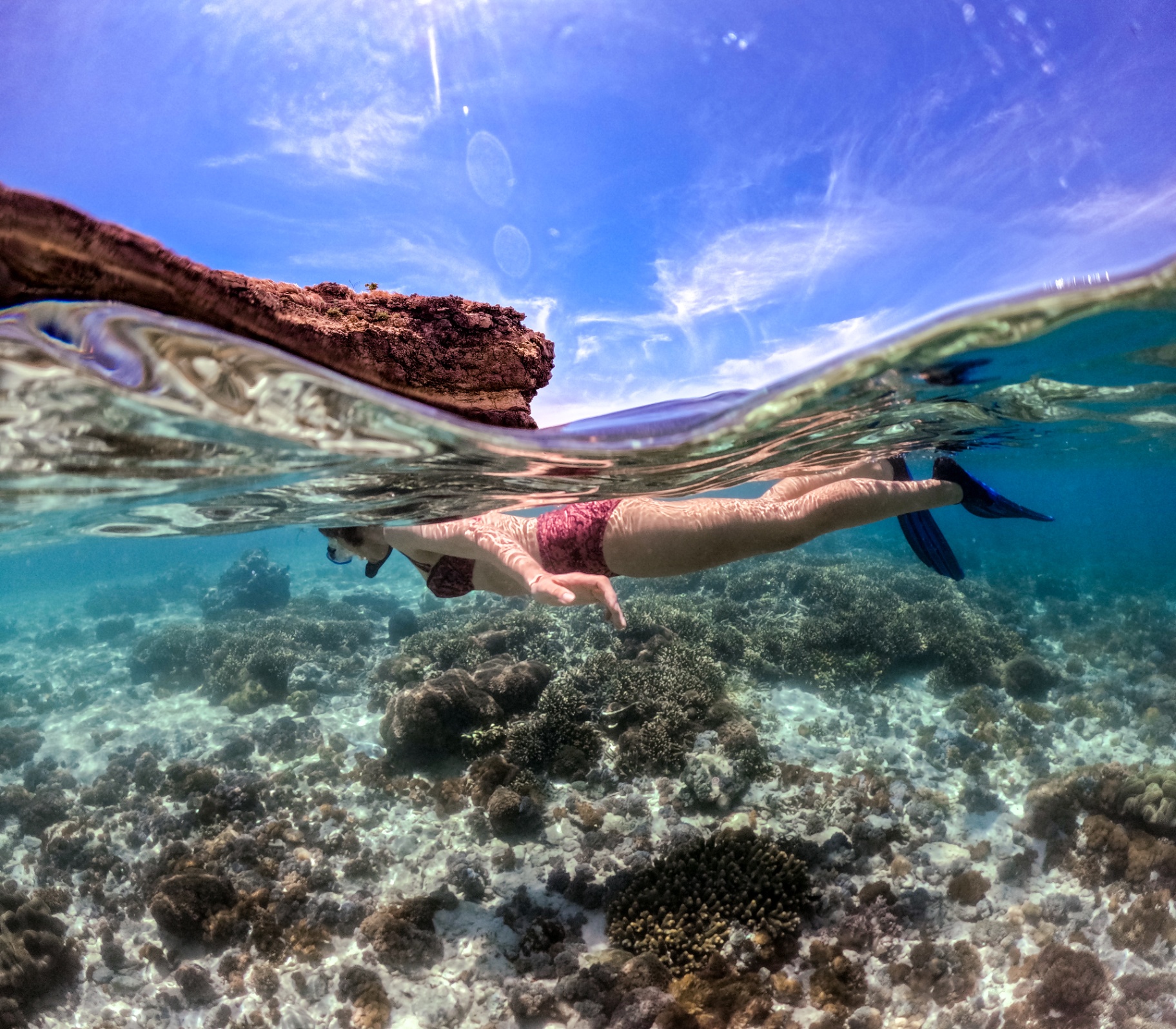 Split-level photo of a snorkeller gliding over shallow Raja Ampat coral with karst island and blue sky above