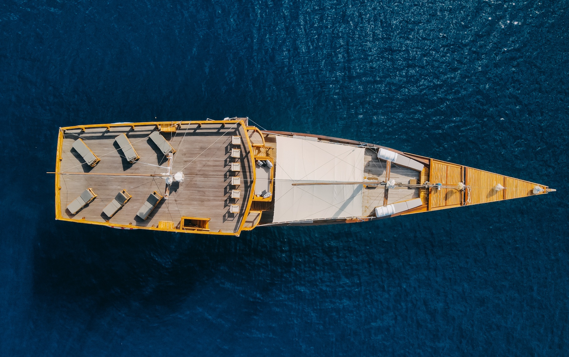 Top-down aerial of Andamari's full deck with sun loungers, shaded lounge and pointed bow on deep blue water