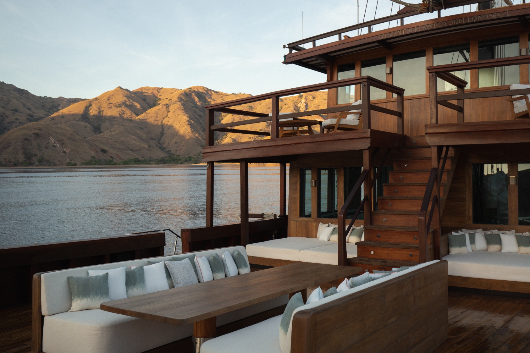 Aft deck lounge on Vela at golden hour with white cushions and daybeds and warm-lit Komodo hills across the water