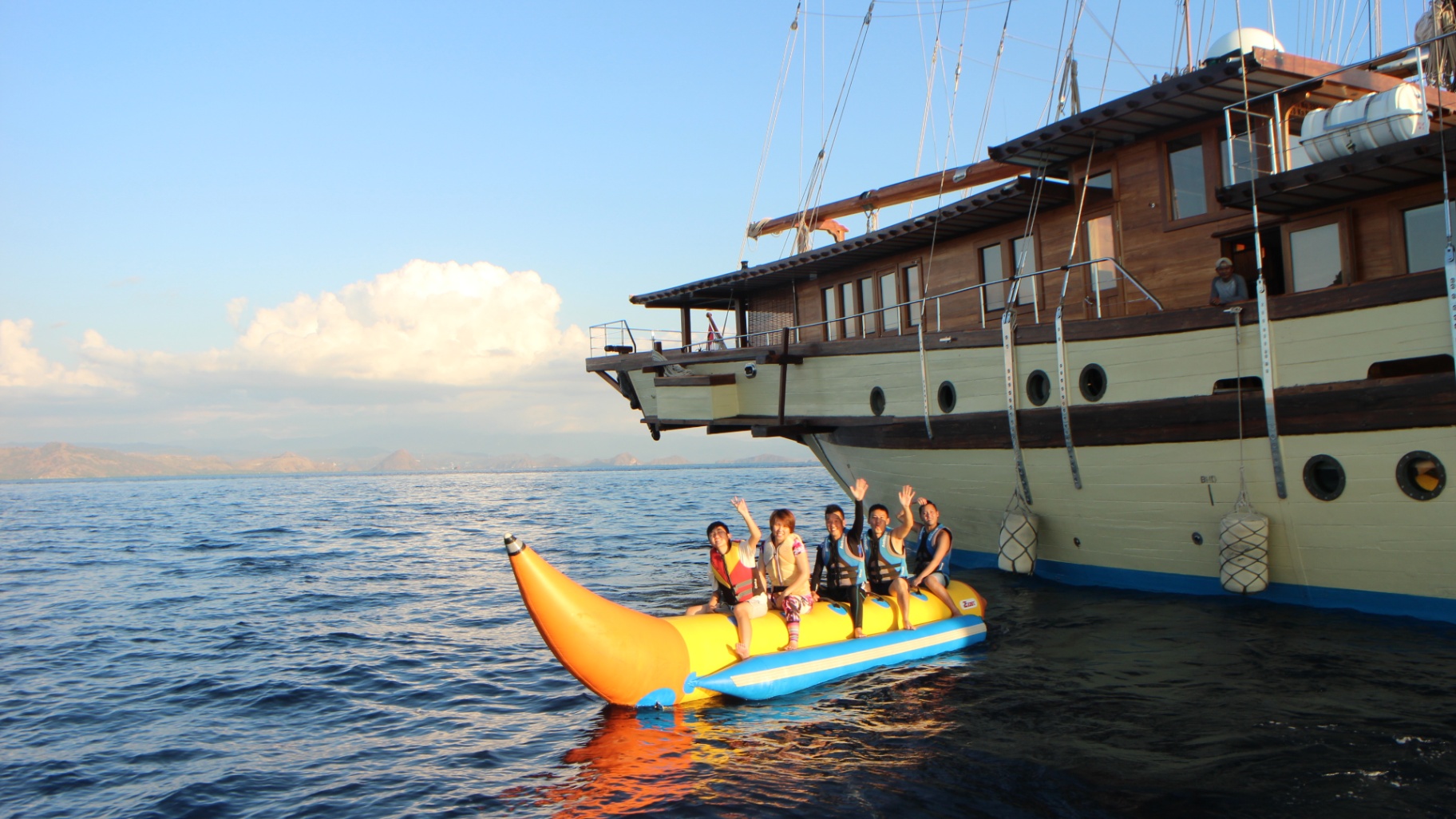 Guests waving from an inflatable banana boat alongside Lamima's hull at sunset