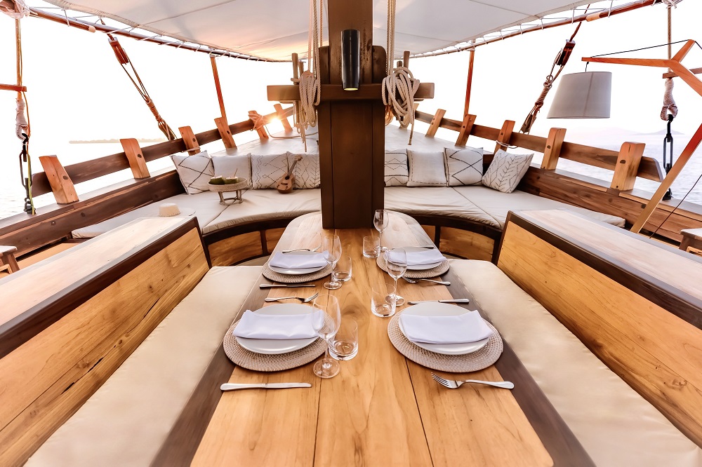 Overhead view down Senja's foredeck dining table with four place settings, wine glasses and cushioned bench seating