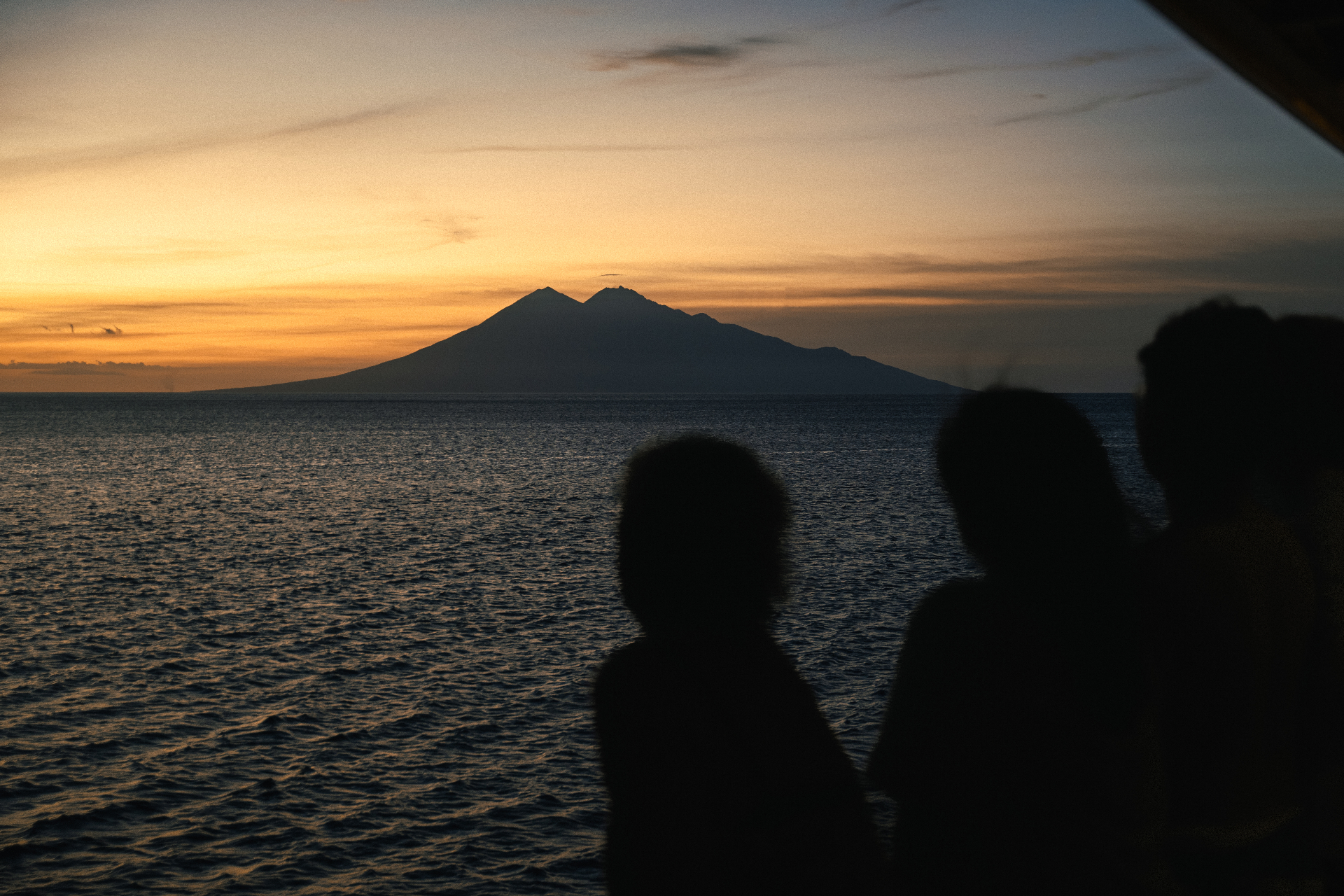 Guests watching sunset from Mischief with a volcanic island silhouetted across the horizon