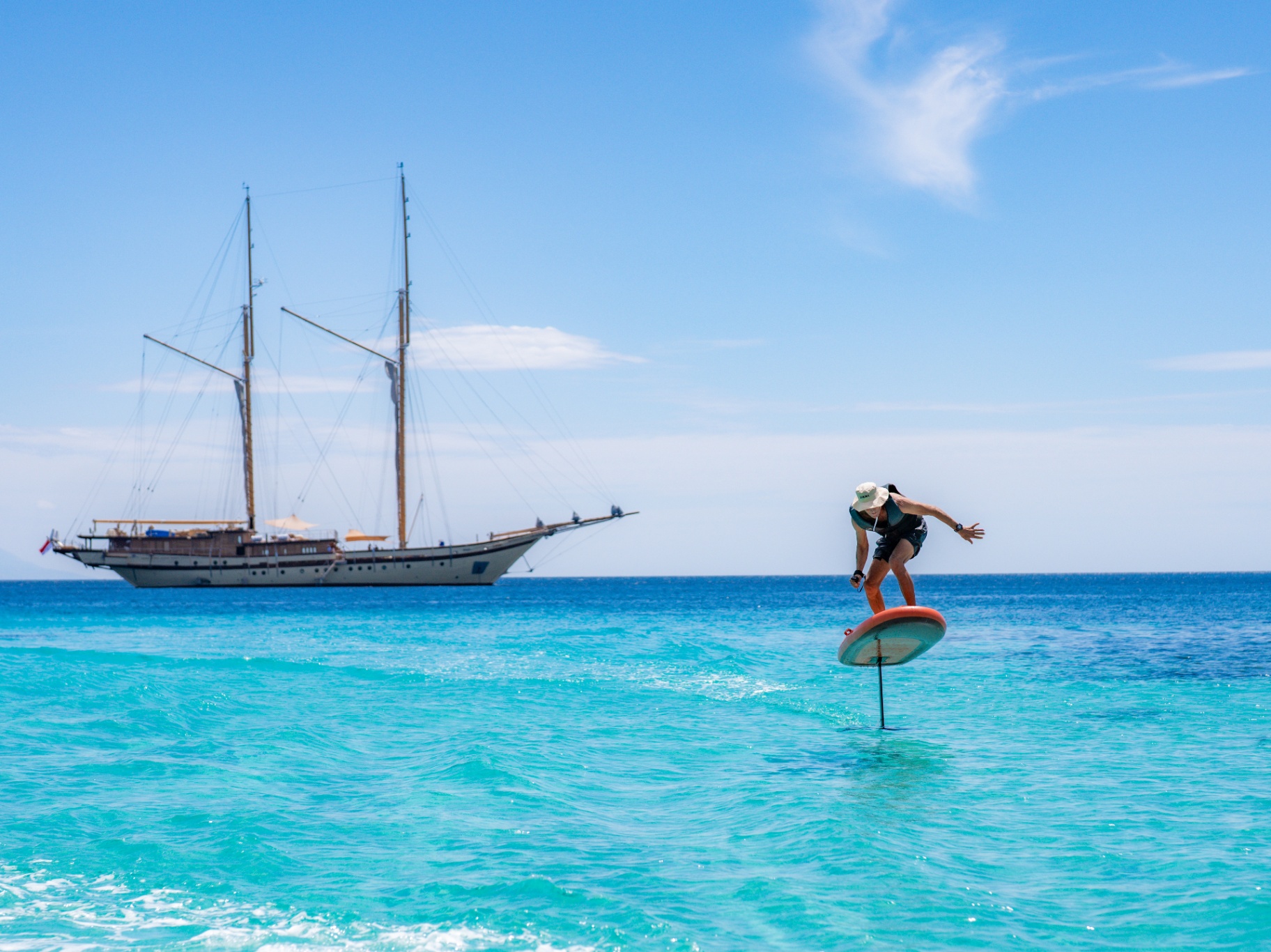 Hydrofoil rider lifting above turquoise water with Lamima at anchor behind