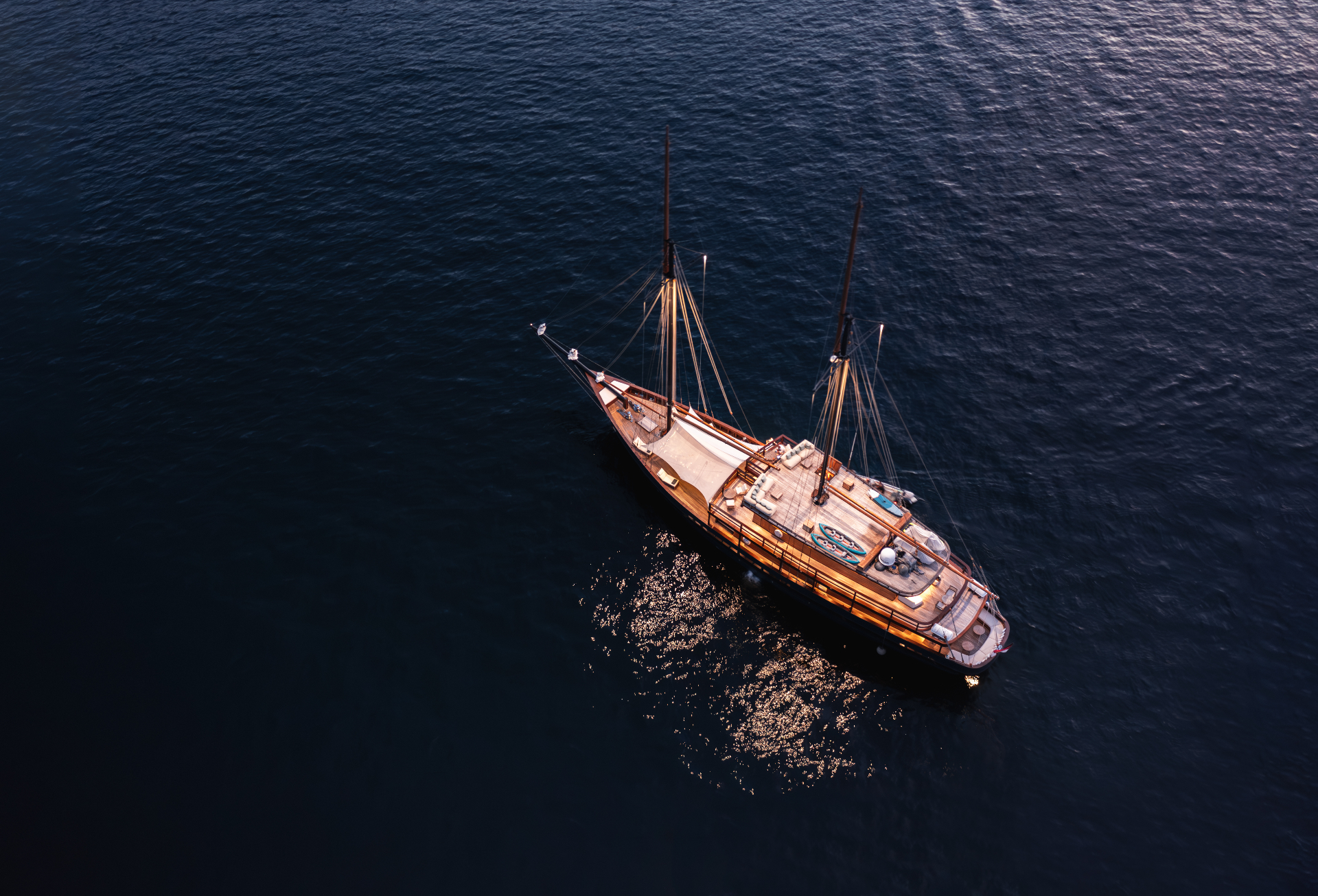 Aerial view of Vela anchored on dark blue water at dusk with warm deck lighting glowing