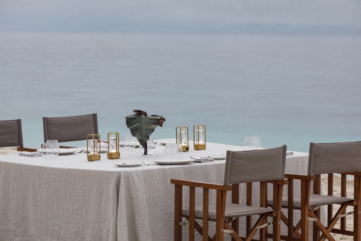 Private beach dinner setup with linen-draped table, lanterns and directors' chairs at dusk