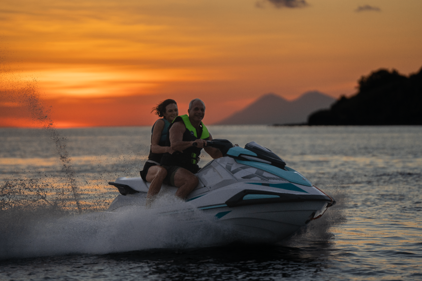 Couple riding a jetski across a calm sea at sunset with a distant volcano silhouetted on the horizon