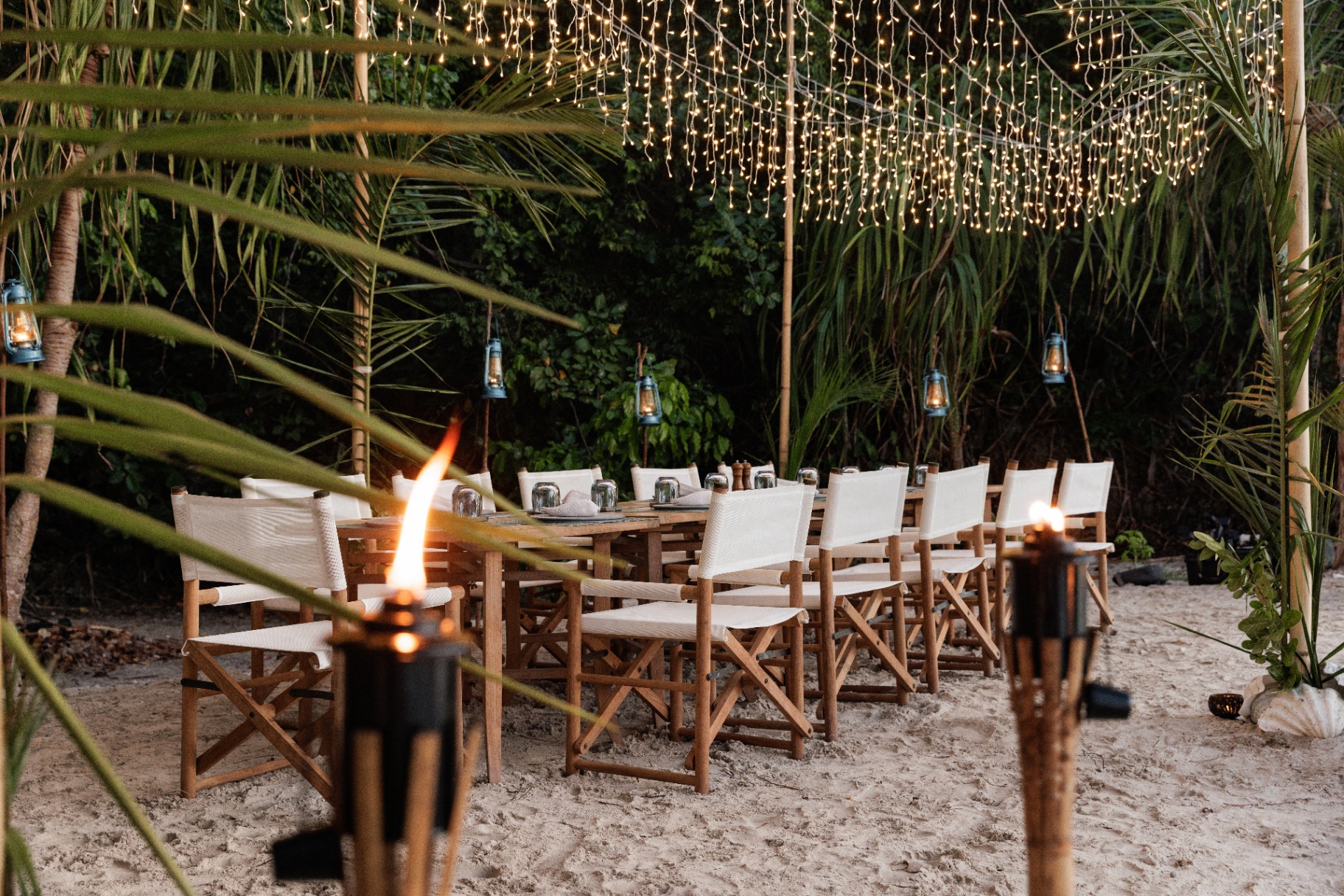 Private beach dinner for Vela guests with a long teak table, director's chairs, hanging fairy lights, lanterns and bamboo torches under palm fronds