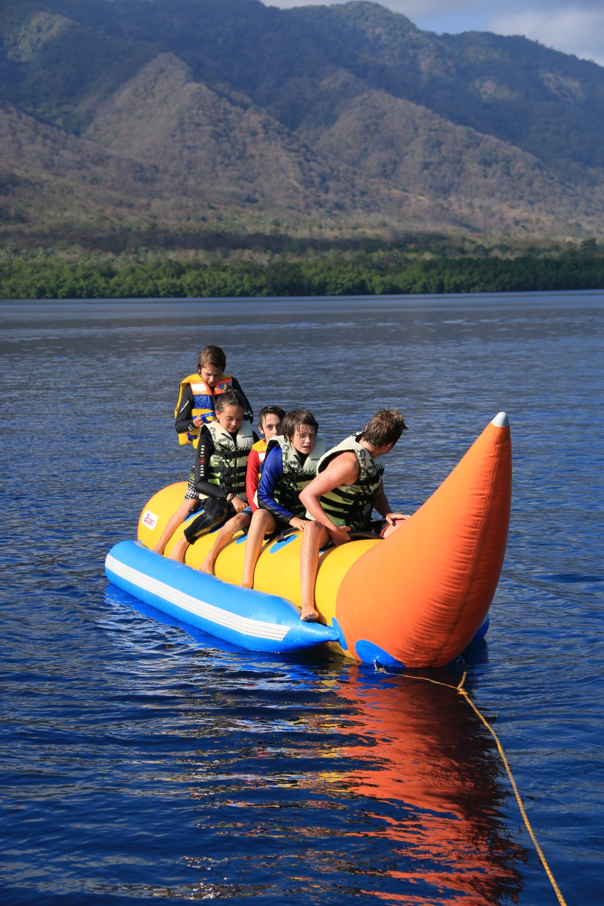 Group of kids in life jackets riding an inflatable banana boat with jungle hills behind