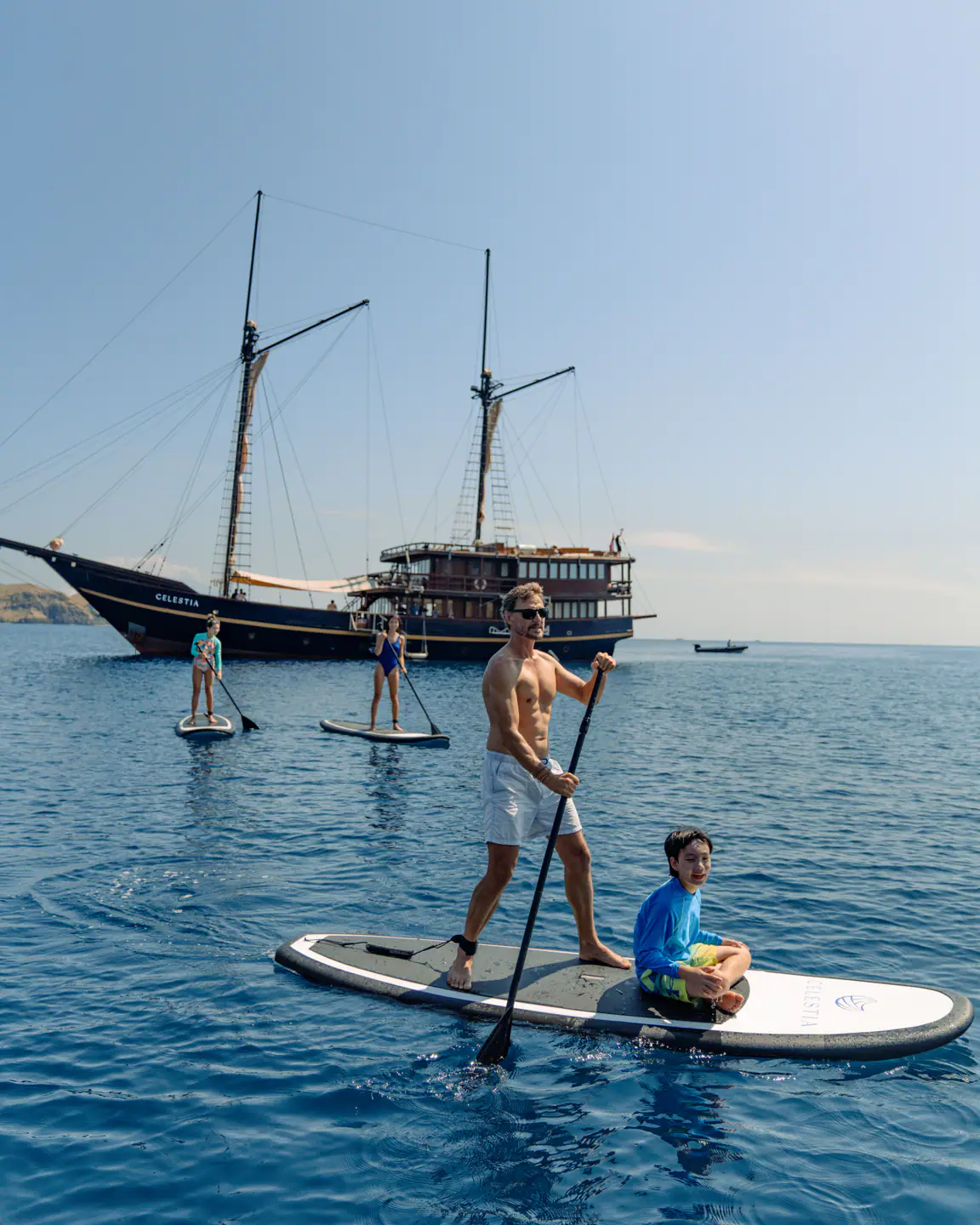 Family paddleboarding beside Celestia phinisi at anchor in Raja Ampat