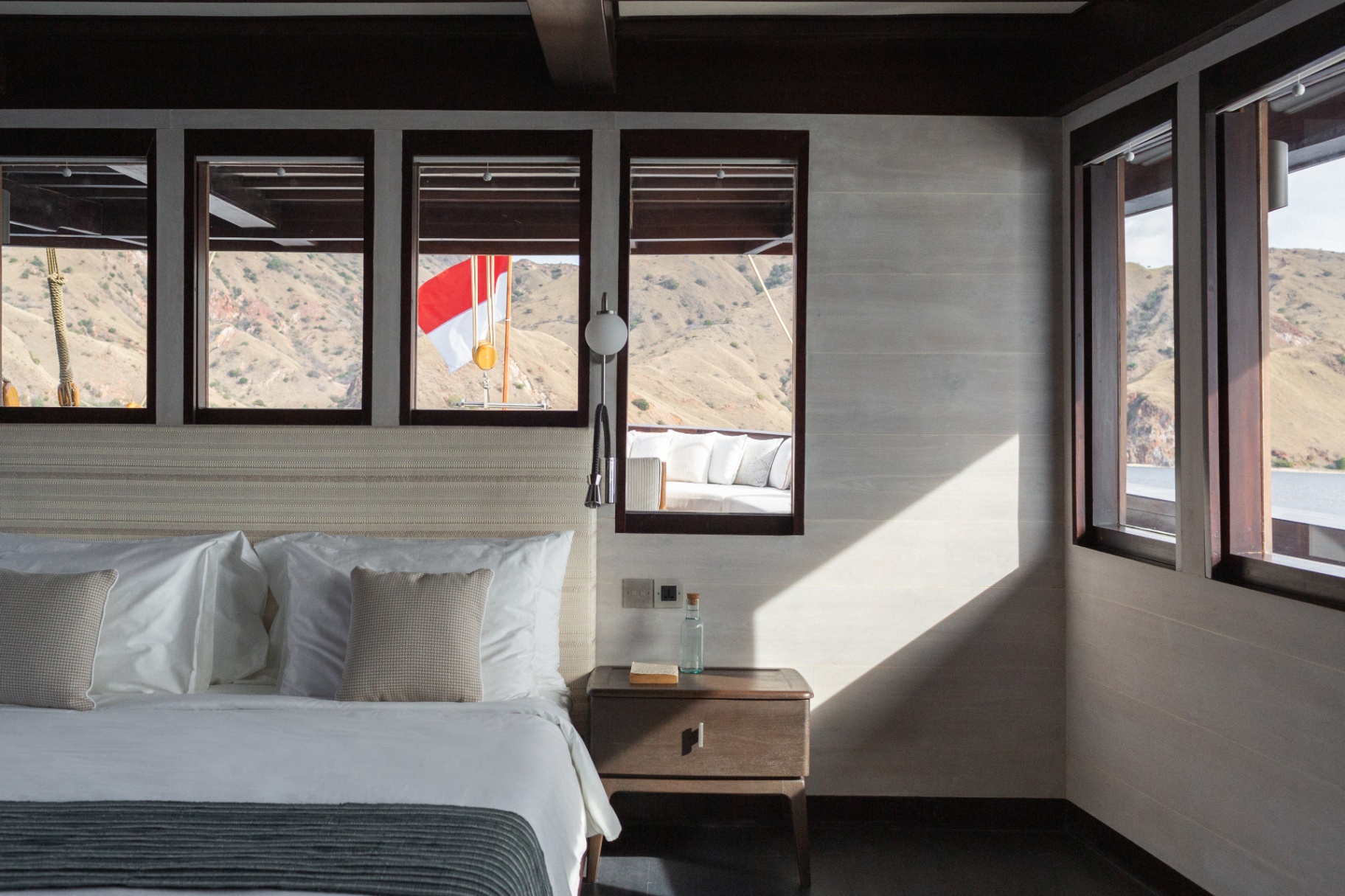 Master cabin on Vela with king bed, linen headboard and picture windows opening to Komodo hills and the Indonesian flag
