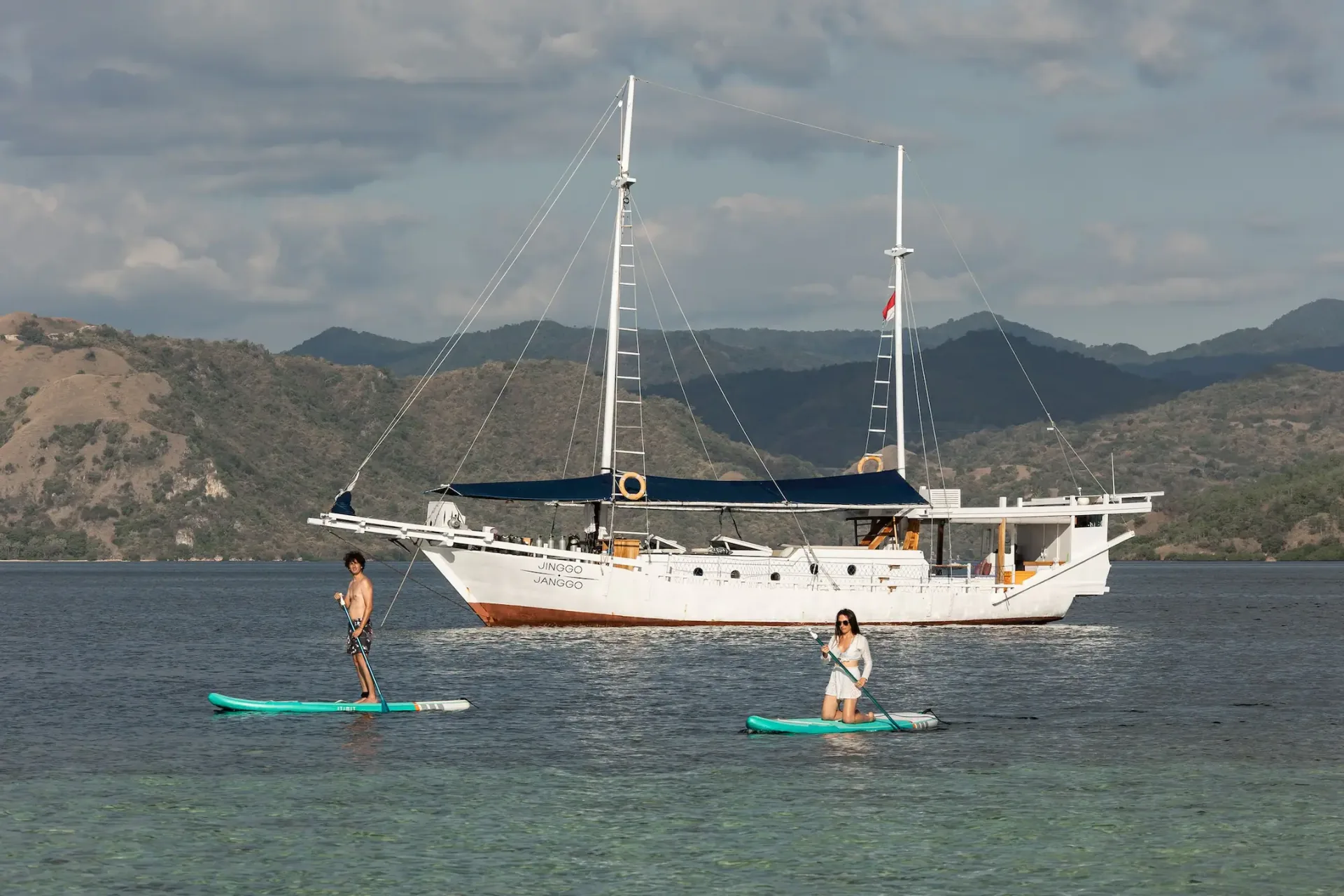 Two guests paddleboarding on calm water beside Jinggo Janggo with Komodo hills behind