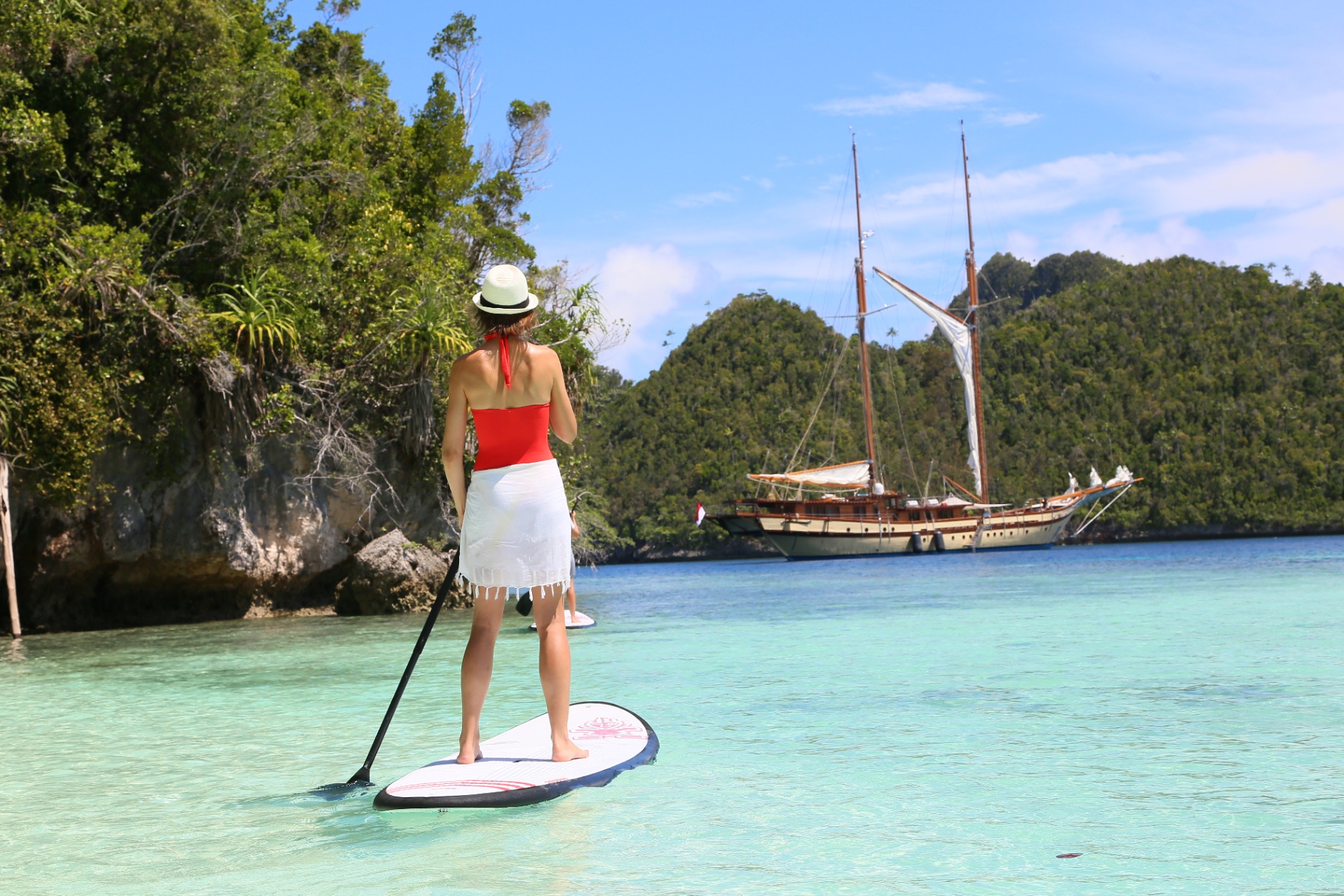 Guest paddleboarding on glassy turquoise water toward Lamima anchored in a Raja Ampat karst lagoon