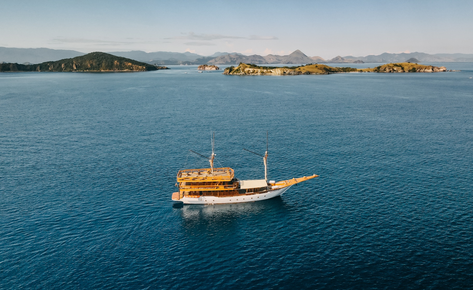 Aerial of Andamari phinisi at rest on glassy blue water with green islands and mountains behind