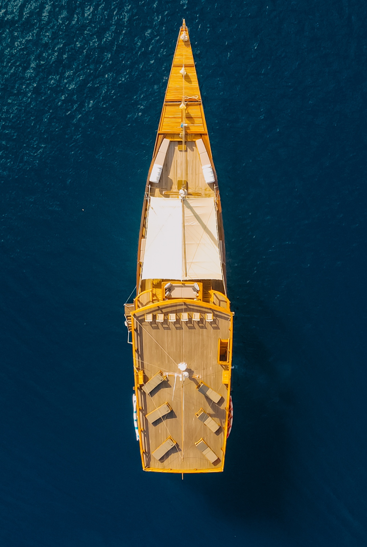 Top-down aerial of Andamari showing the teak upper deck with sun loungers, sun sail and pointed bow on deep blue water