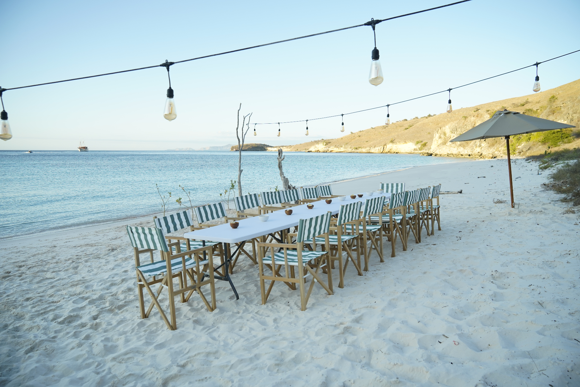 Long beach dinner table set with green striped chairs and festoon lights on a white-sand Raja Ampat beach