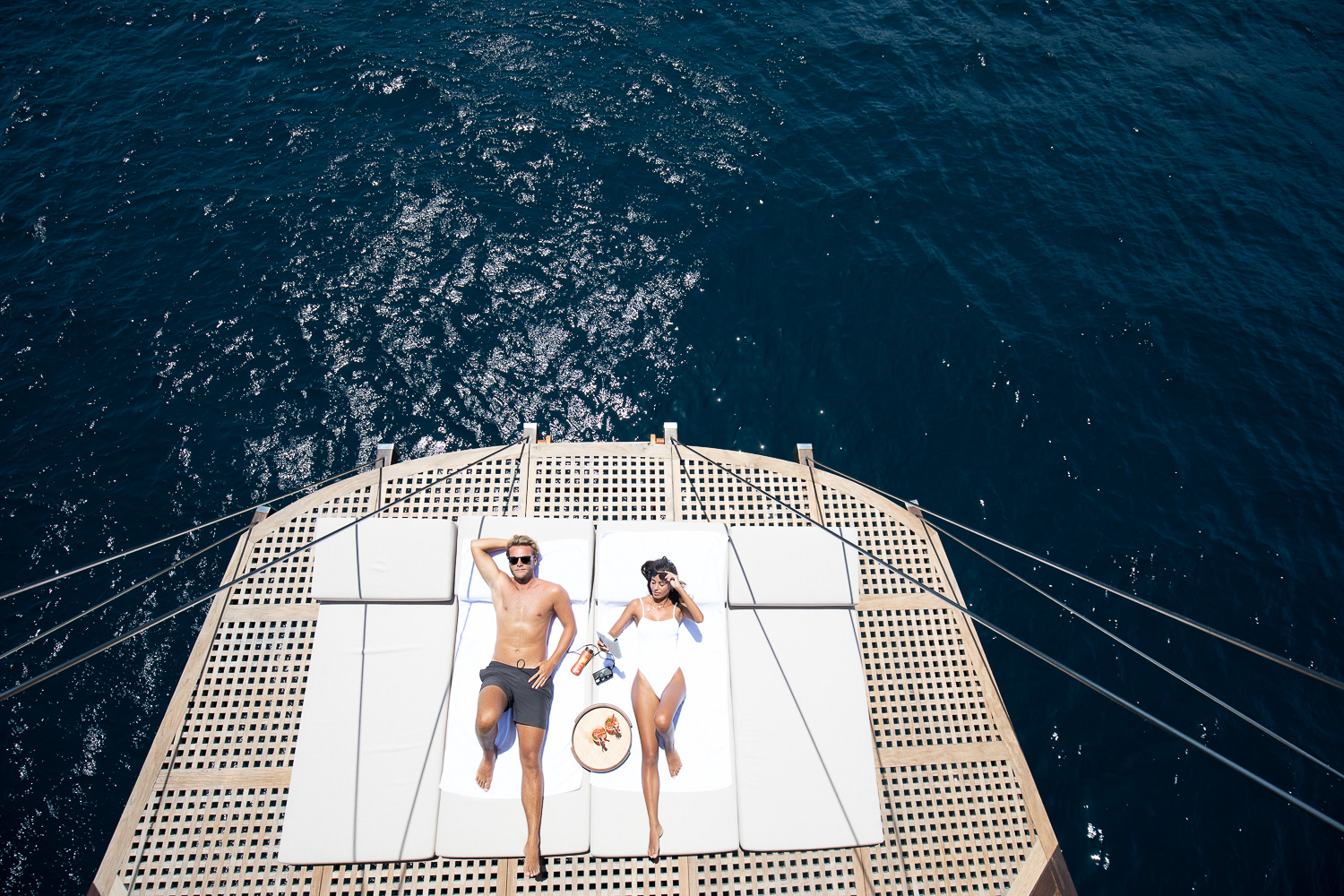 Aerial view of a couple sunbathing on bowsprit loungers over deep-blue water