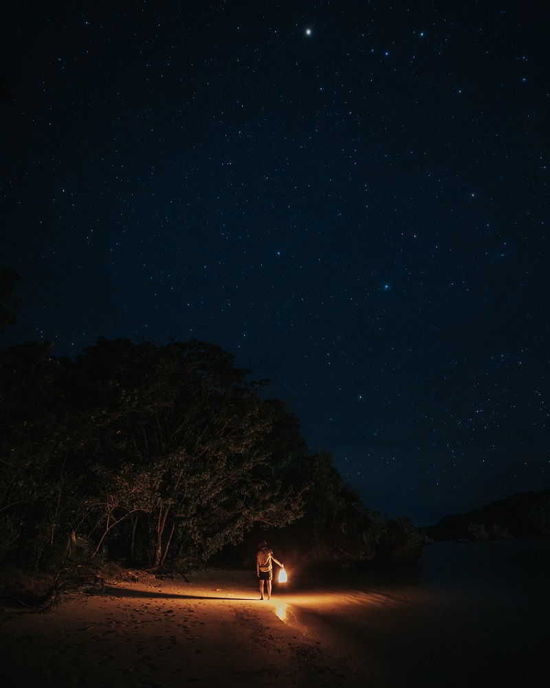 Guest walking a Raja Ampat beach at night with a lantern under a sky full of stars