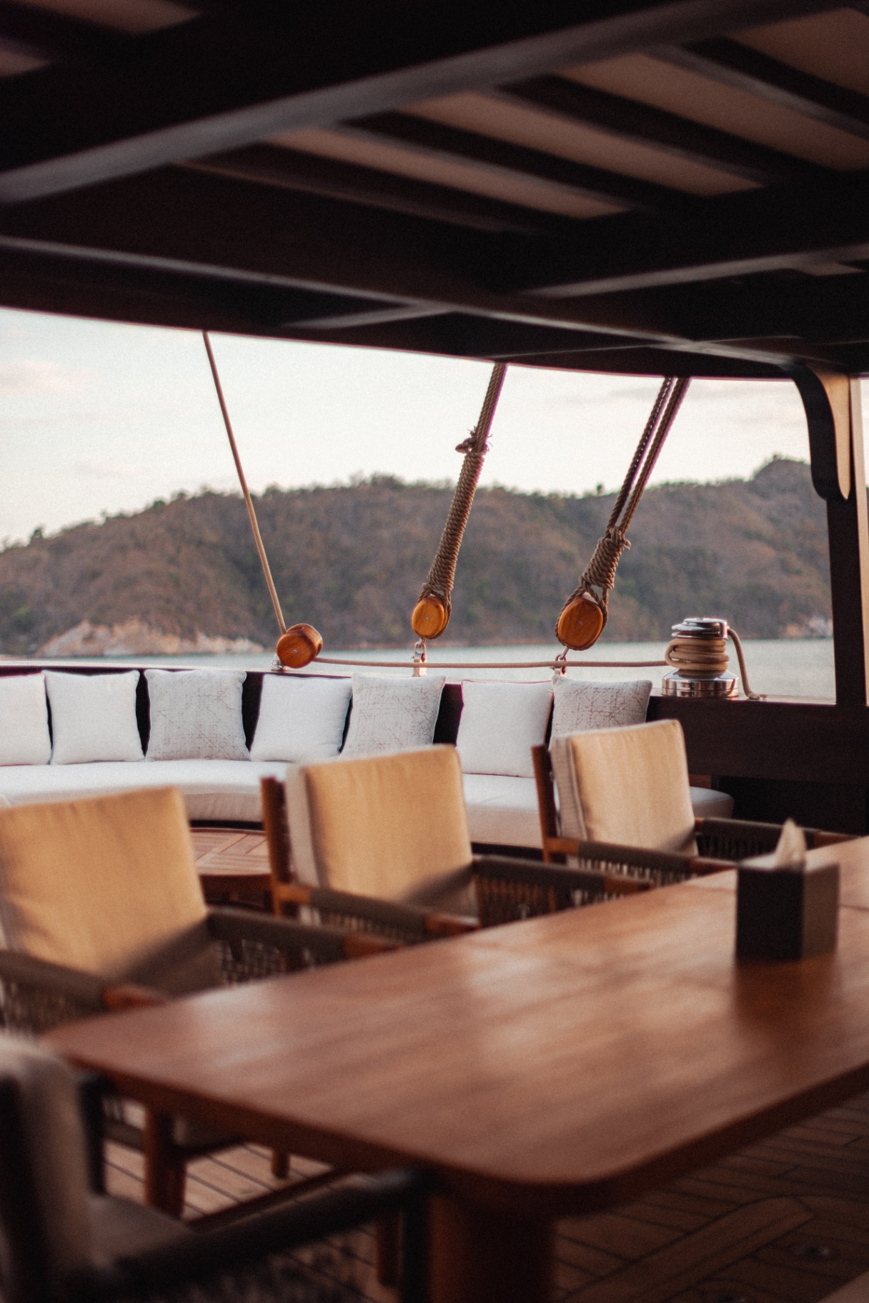 Aft deck lounge on Vela with white cushions, wooden blocks and rope rigging framing a view of green Komodo hills