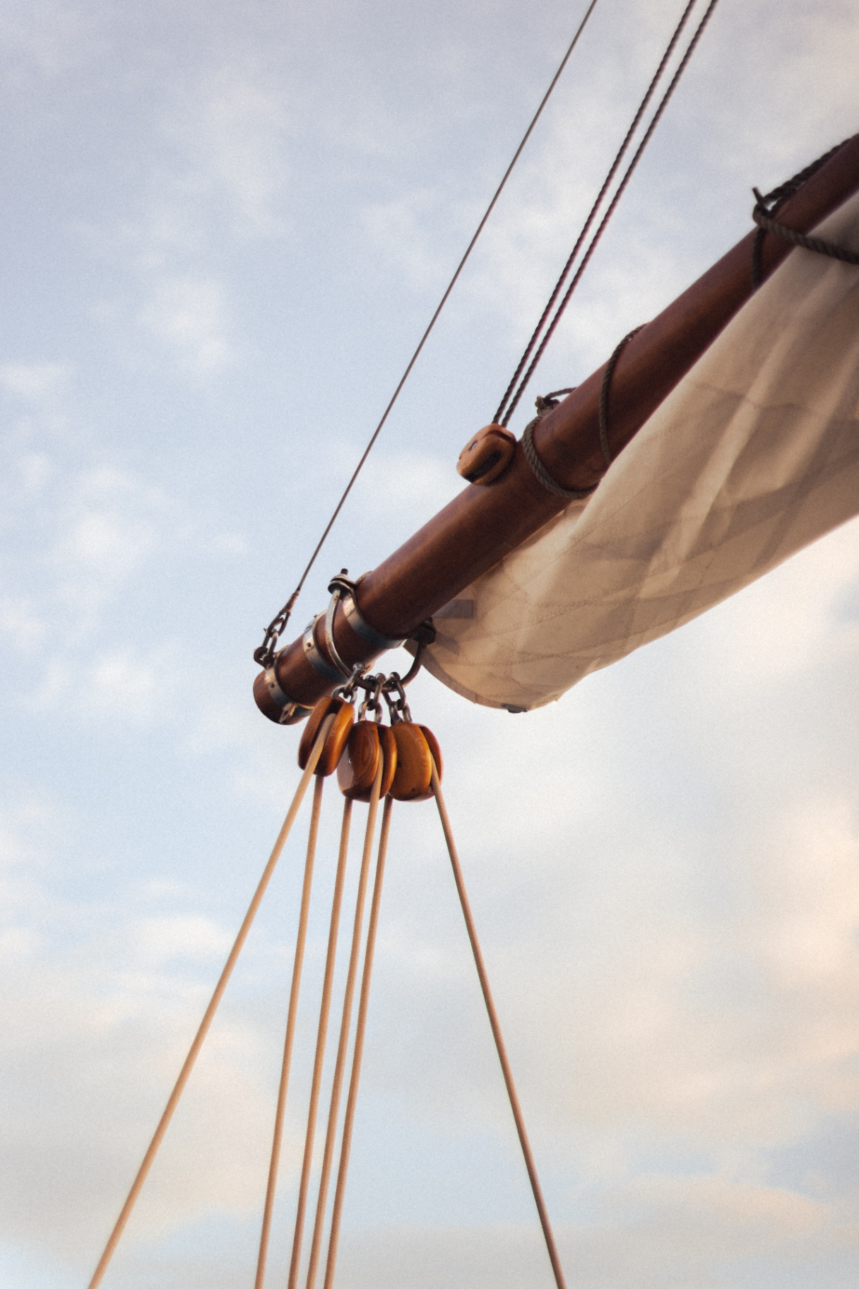 Detail of Vela's varnished boom, wooden pulley blocks and rope tackle against a soft blue sky