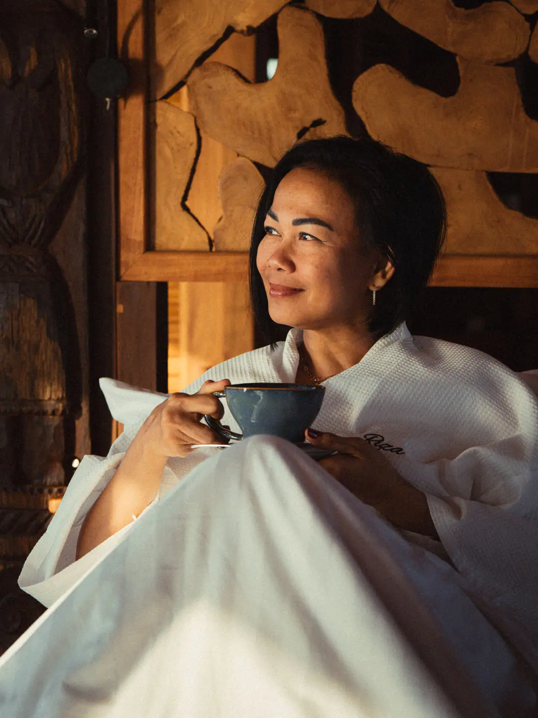 Guest in white robe smiling with a cup of coffee in warm cabin light on The Oracle