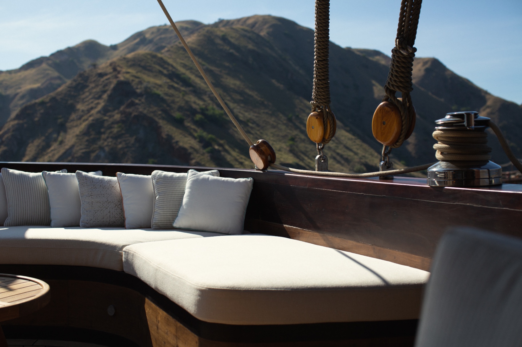 Curved deck banquette with linen cushions, wooden blocks and winch, framed by dramatic Komodo hills