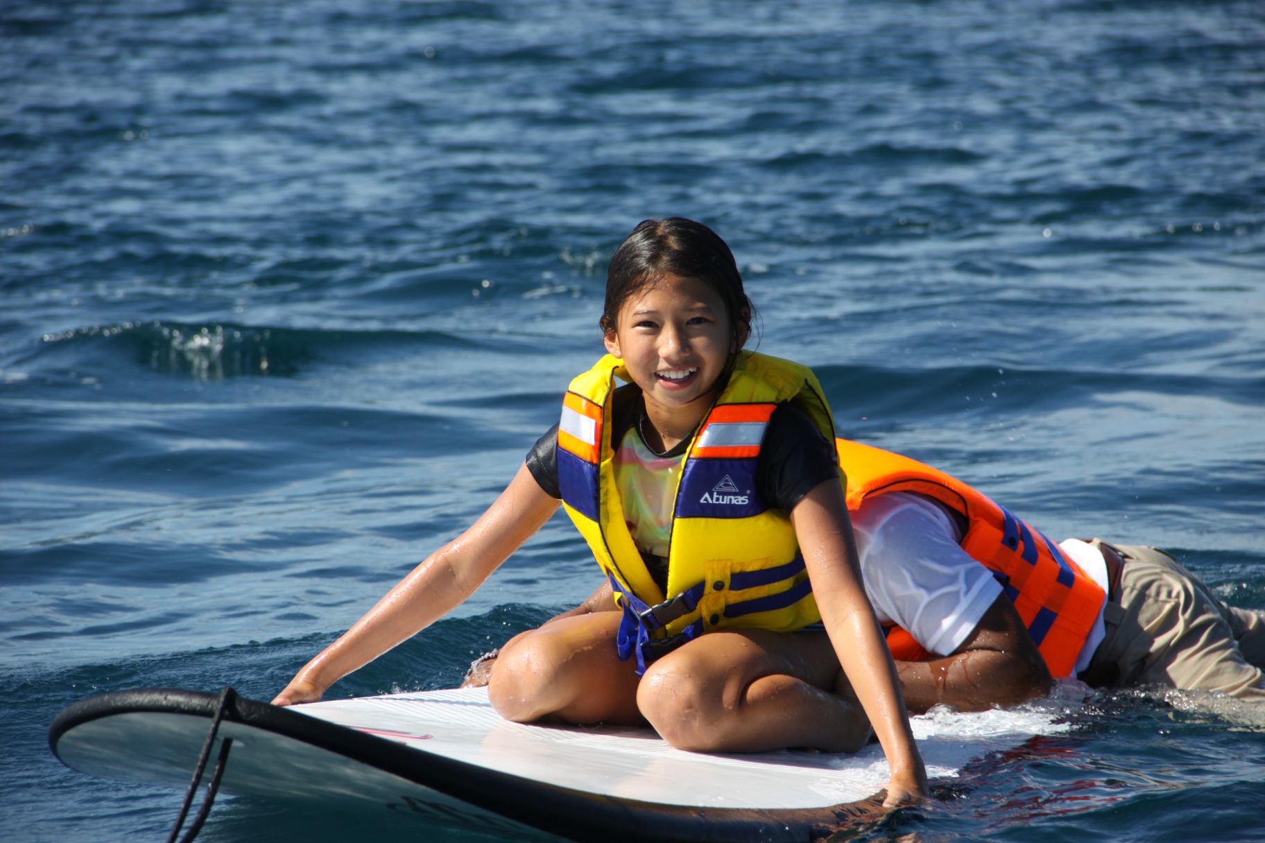 Smiling child in a life jacket sitting on a paddleboard being towed across calm blue water