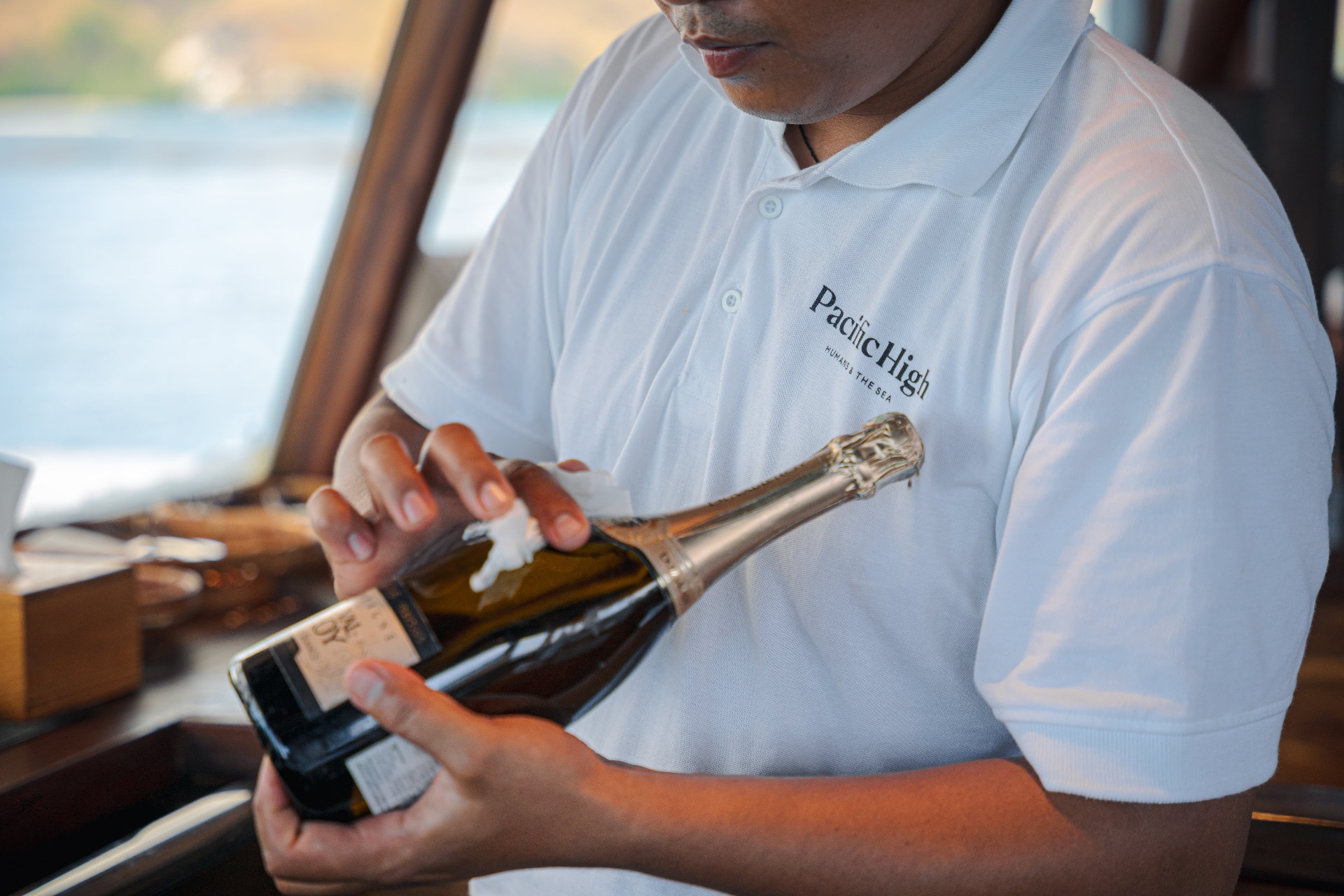 Crew member in a Pacific High polo opening a bottle of champagne on Nataraja