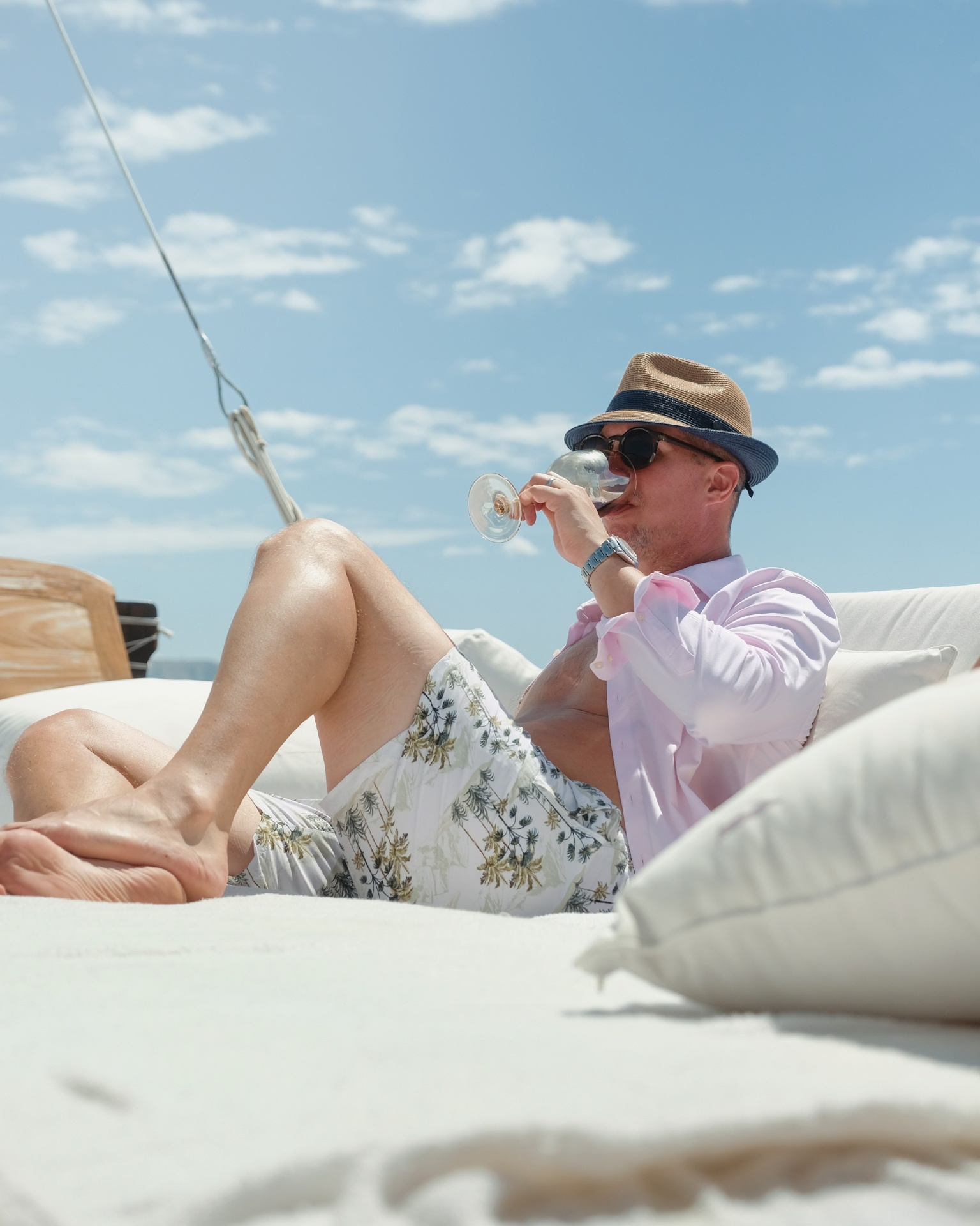 Guest in straw hat and sunglasses relaxing on Samara II's forward sun cushions with a glass of wine