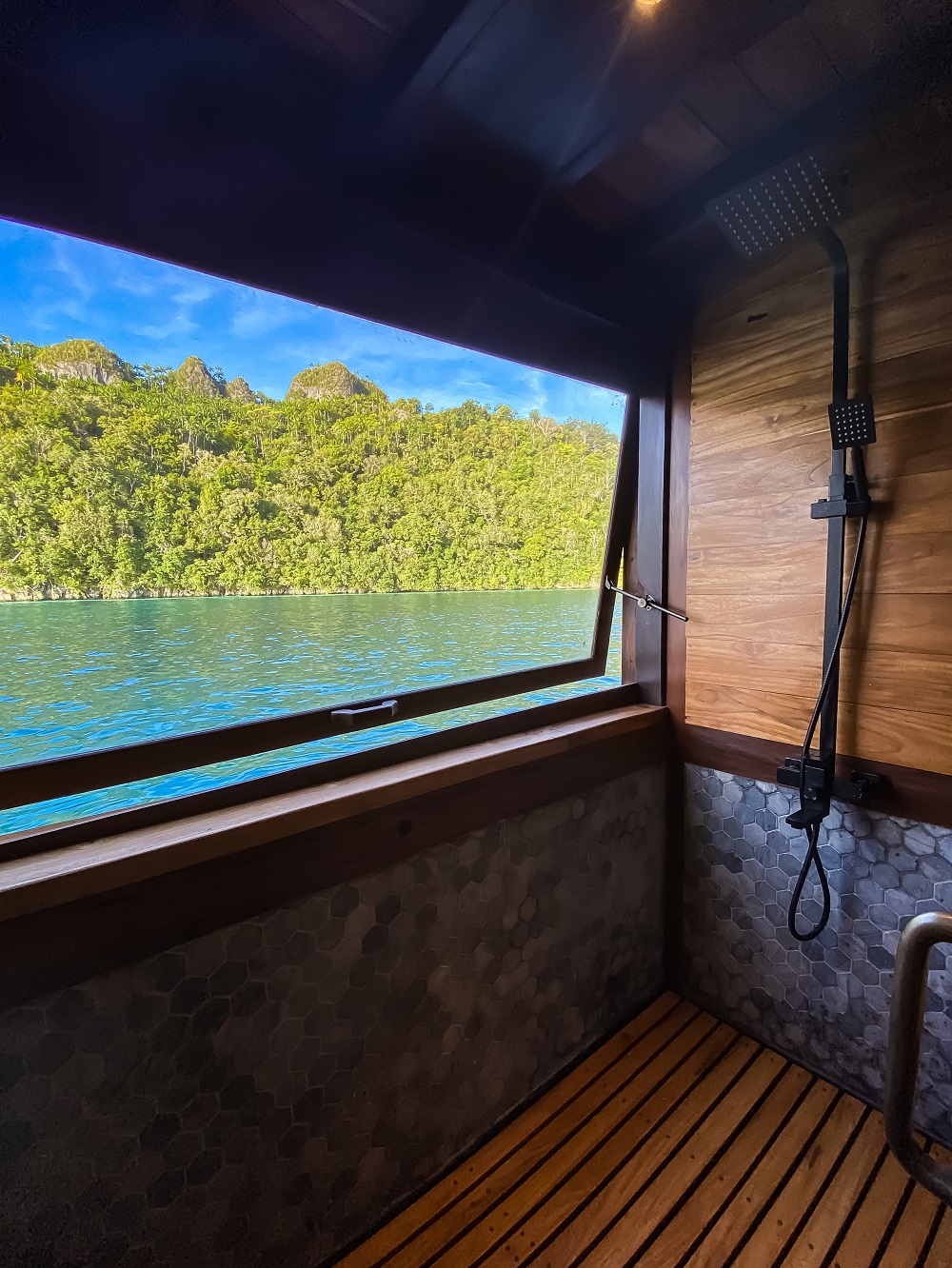 Portrait view of Senja's shower with rain head, teak deck and jungle island beyond the window
