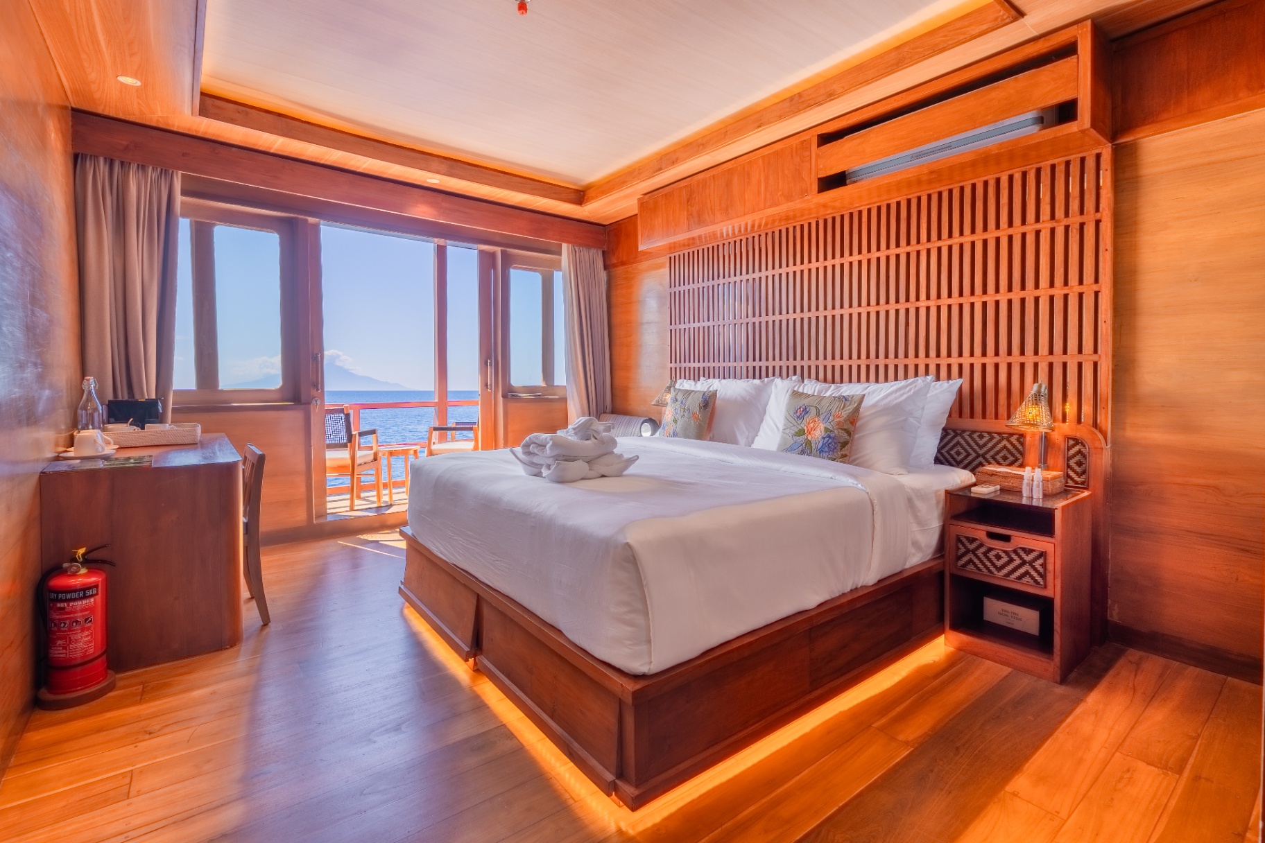 Deluxe cabin on Sanctuary with king bed, slatted teak headboard and sliding doors to a private sea-view balcony at sunset