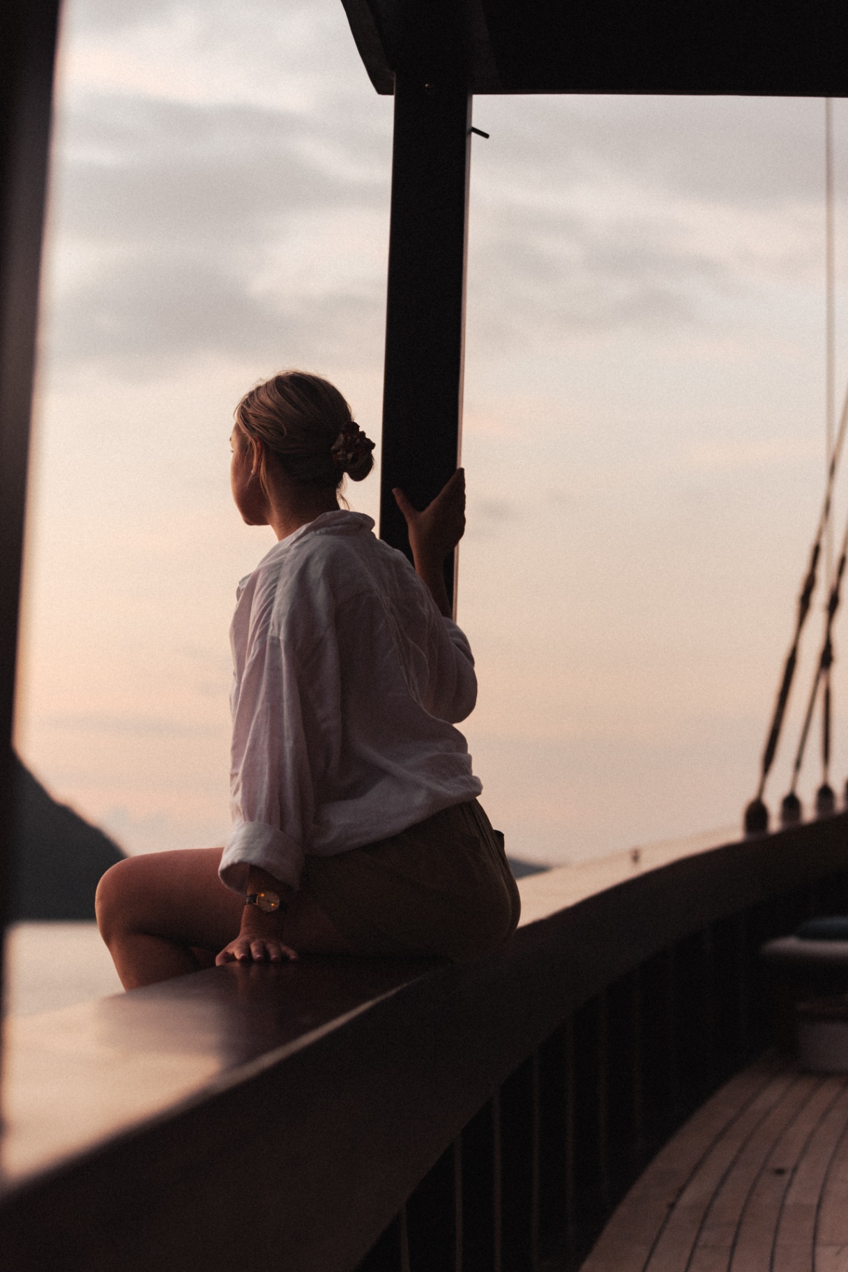 Guest in a white shirt sitting on Vela's teak rail watching a soft pink sunset over the sea