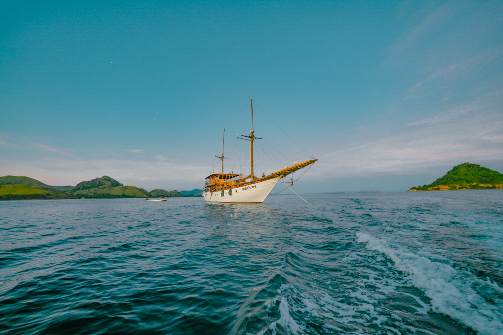 Adishree phinisi at anchor at dusk with soft pink sky and green islands surrounding the yacht